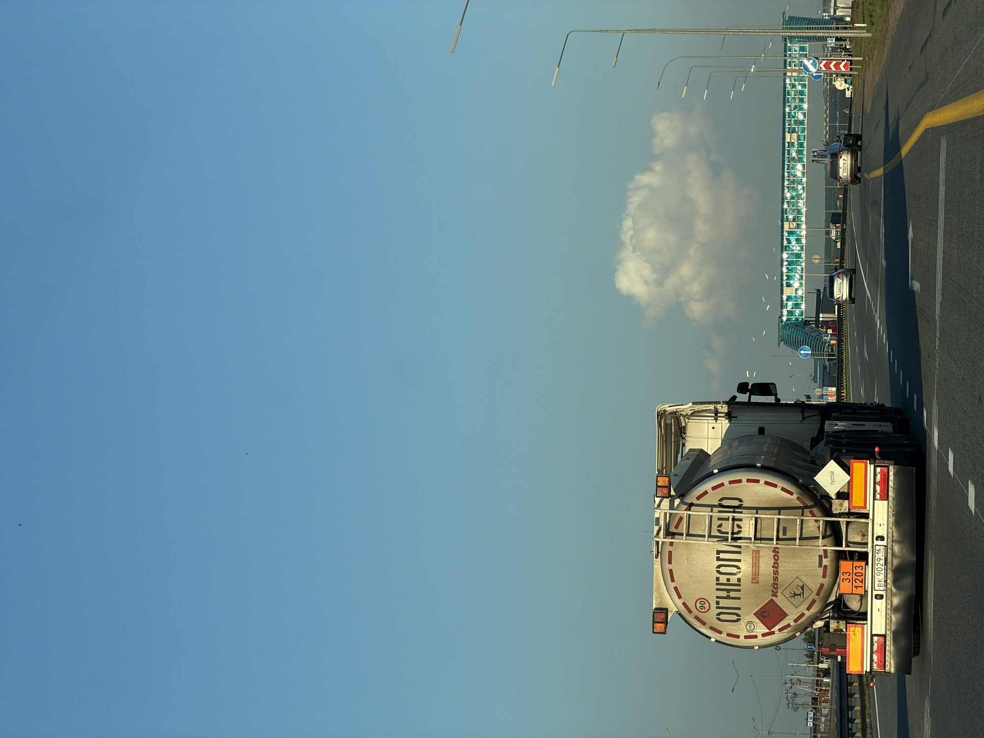 The image depicts a highway scene in Kulaevo, Russia, under a clear blue sky with a single large white cloud. A white tanker truck, identified by the Cyrillic text "ОГНЕОПАСНО" (Flammable) and hazard symbols, is in the foreground, driving on the right side of the road. The truck has a ladder on its side and license plates visible. Several other vehicles, including cars and a smaller truck, are also present on the highway. In the background, a modern pedestrian bridge with a green, translucent roof spans the highway. Streetlights line the sides of the road, and vegetation is visible beyond the highway on either side. The overall impression is of a typical day on a Russian highway, with the tanker truck being the most prominent subject.