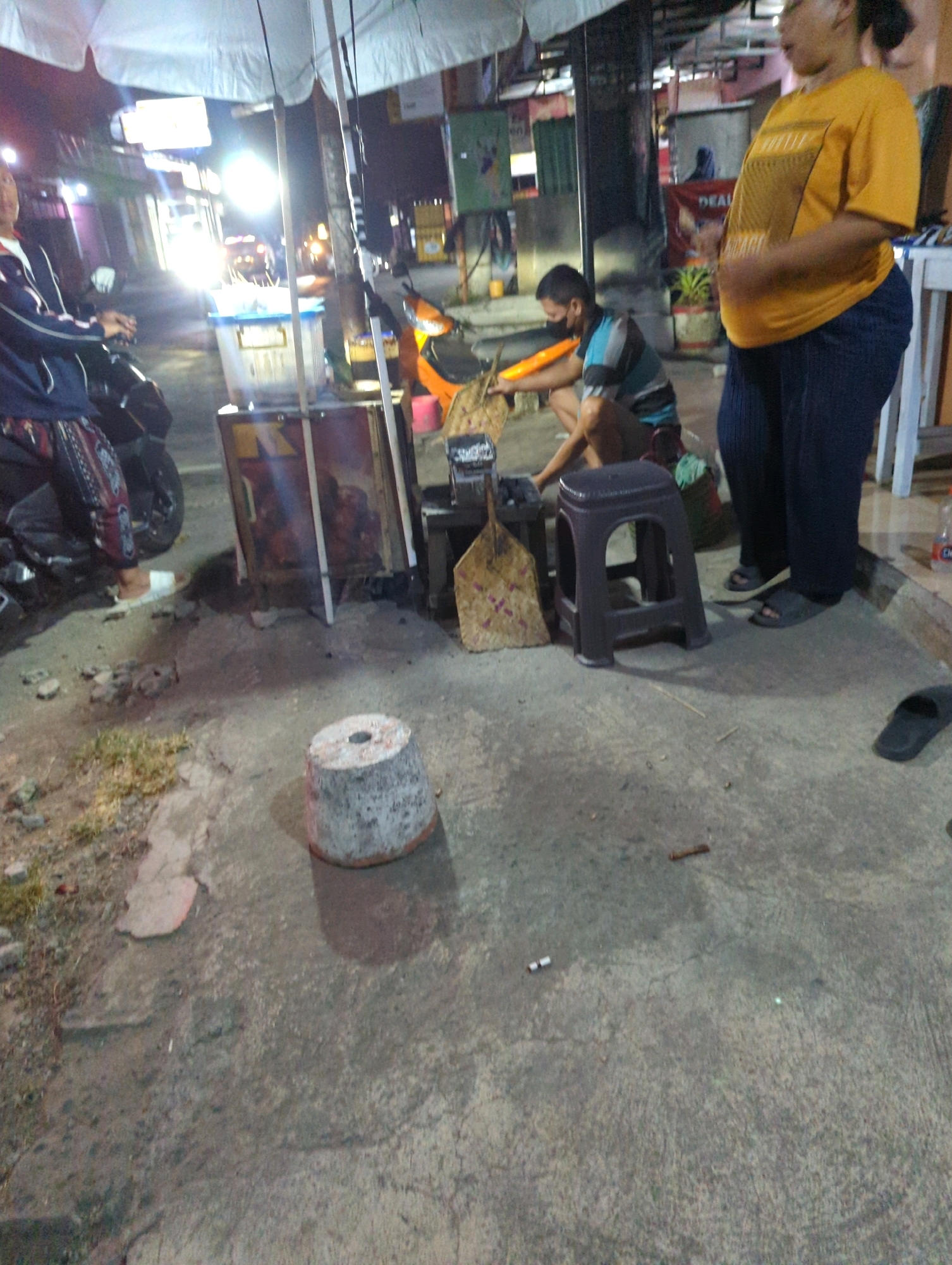 In Kacangan, Indonesia, a nighttime street scene unfolds under a large white umbrella that provides shade for a food stall. The ground is paved with concrete, showing signs of wear and tear with cracks and scattered debris. To the left, a person wearing a dark blue jacket with white stripes and a patterned fabric skirt is partially visible, seated on a motorcycle. In the center, a man wearing a blue and white striped shirt and dark pants is crouched down, attending to a small charcoal grill. He wears a dark face mask. A dark plastic stool sits to his right. To the right, a woman with dark hair, wearing a bright yellow t-shirt with a graphic print and dark pants, stands and observes the scene. Behind the food stall, various items are visible, including a blue cooler and what appears to be a red and yellow striped object, possibly a sign. An orange scooter is parked nearby. The background depicts a street with buildings and illuminated signs, suggesting a commercial or residential area. The lighting is dim, with bright lights from the street illuminating parts of the scene.