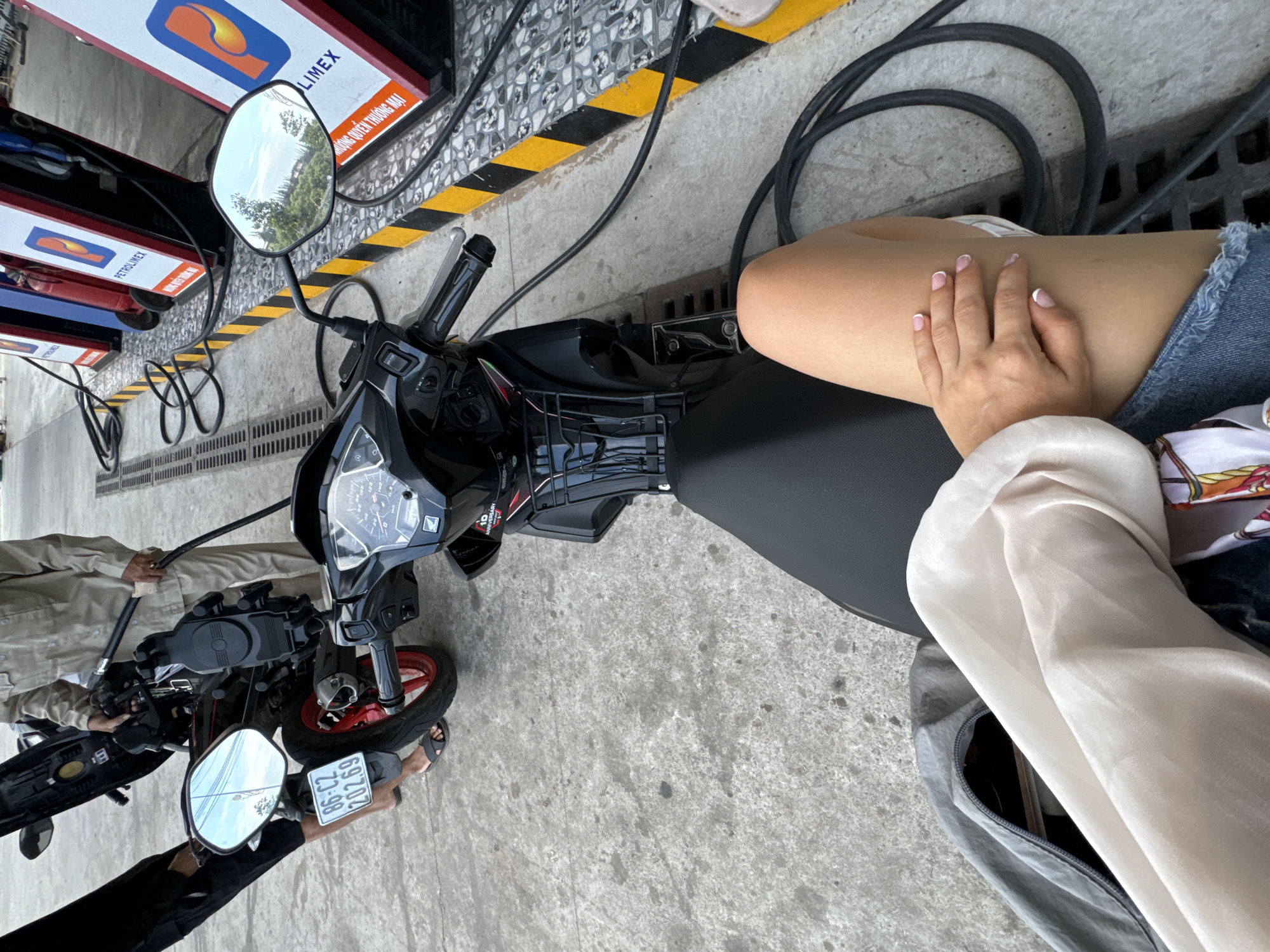 The image depicts a scene at a Petrolimex gas station, specifically located in Phu Thuy Ward, Vietnam.
Foreground: A woman is sitting on a black scooter. She is wearing denim shorts and a light-colored blouse. Her hand rests on her thigh. Beside her is a gray bag.
Midground: A black scooter is at a Petrolimex gas pump. To the left, another person wearing light-colored clothing is holding the handlebars of another scooter. The second scooter also has a red rim around the front tire.
Background: The Petrolimex gas pumps are visible with red and blue colors. Black hoses extend from the pumps. The ground is concrete and tiled with a yellow and black safety marking.