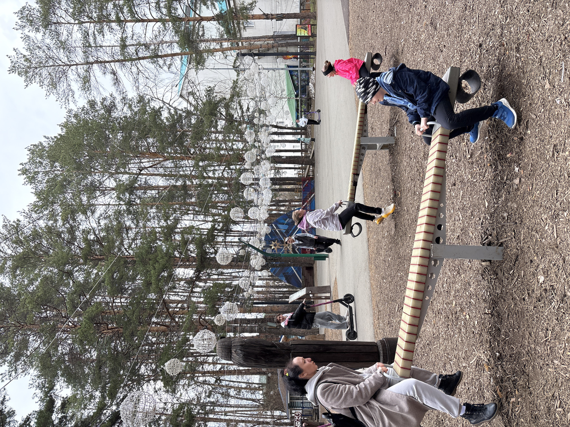 The image shows a public space in Neftekamsk, Russia. It depicts several people engaging in recreational activities. In the foreground, a woman in a light-colored coat and grey pants sits on a seesaw. Further along the path, a person on a scooter and two individuals on another seesaw are visible. A young boy with a helmet and blue shoes is situated at the higher end of the second seesaw. Overhead, a string of decorative spherical lights hangs between tall trees. In the background, there are more people, buildings, and signs. The ground is covered with wood chips or mulch in the immediate vicinity of the seesaws. The weather appears overcast.