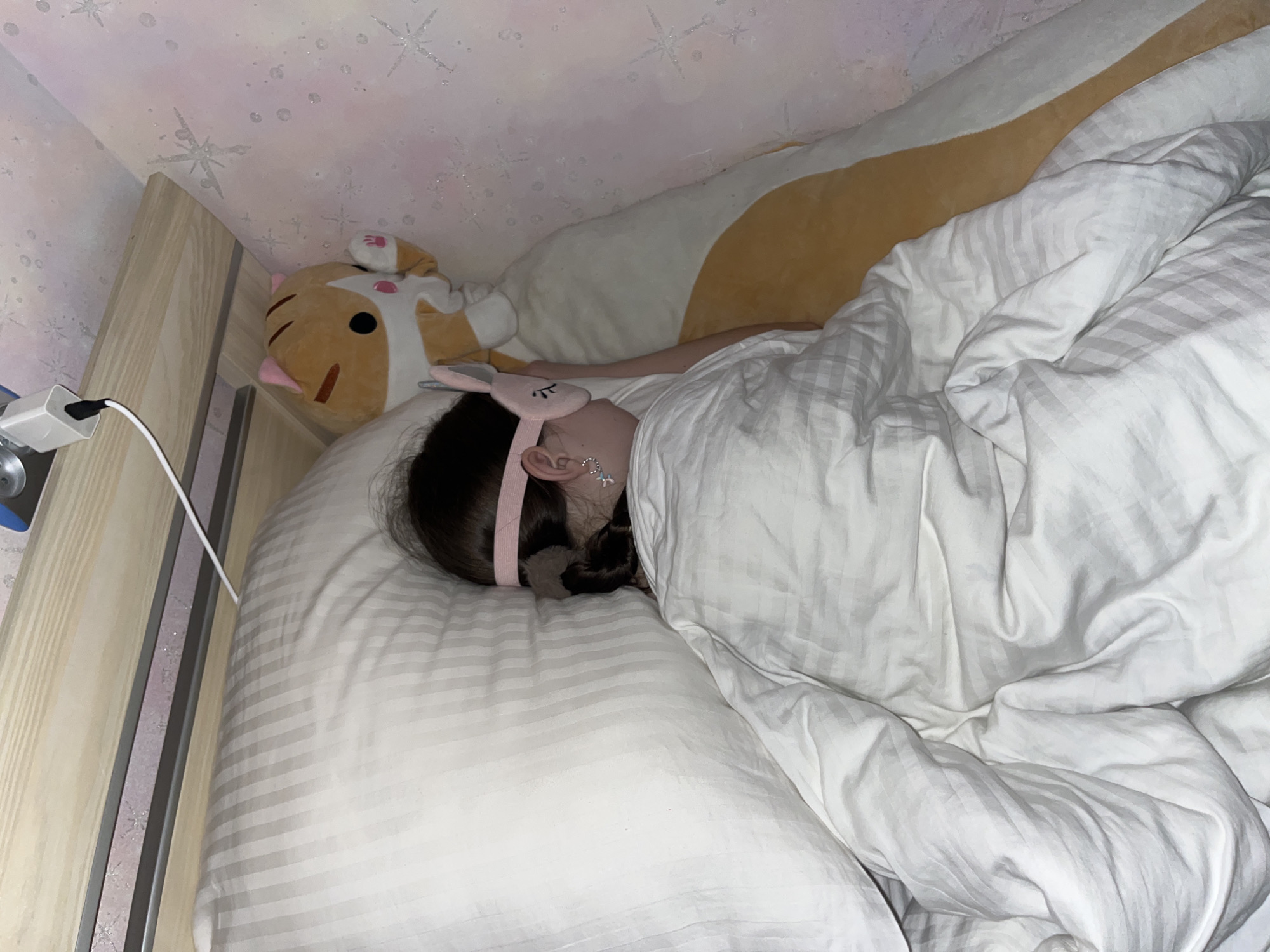 A girl sleeps in a bed. She wears a pink sleep mask with bunny ears. Her black hair is tied back. The bed has striped white sheets. A stuffed animal cat is near her head. A long, yellow stuffed animal is above her. A charger cable is plugged into an outlet on the wooden bed frame. The wall has snowflake wallpaper. The location is unknown, but Chaikovsky, Russia, is provided as the city.
