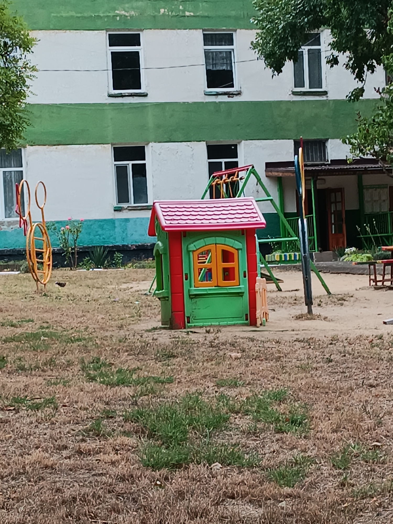 The image depicts a schoolyard in Chișinău, Moldova, featuring a multi-story building with a white facade and green horizontal stripes. Several windows are visible on the building's exterior. In the foreground, a playground area is present, containing a small, brightly colored playhouse with a red roof and green and red walls. Adjacent to the playhouse is a swing set with green metal A-frames and colorful swing seats. A yellow circular toy, possibly a ring toss game, is also located on the playground. The ground is covered with dry, brown grass and patches of greener vegetation, suggesting a summer season. The overall environment appears to be an outdoor recreational space associated with an educational institution.