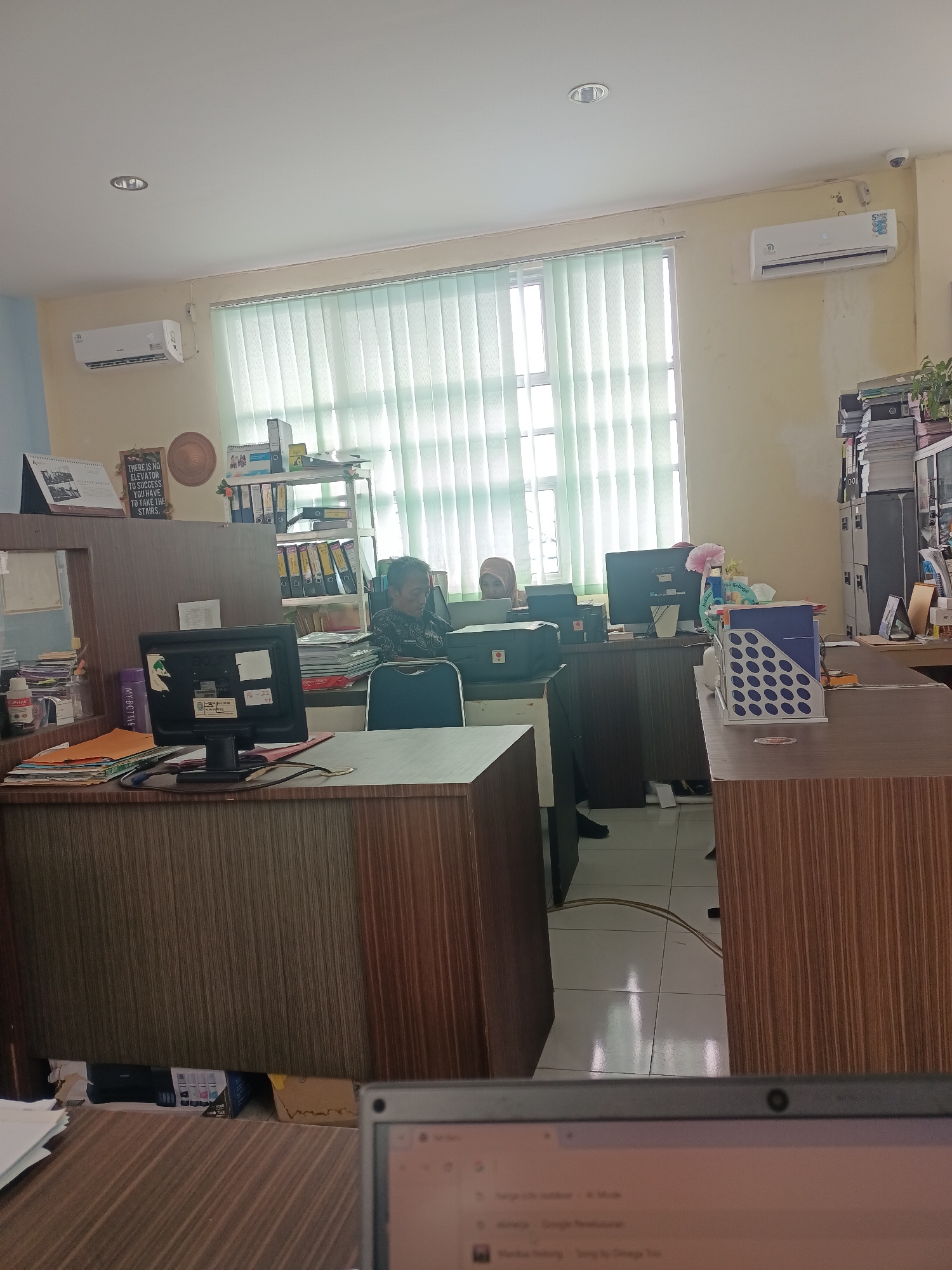 This is an indoor scene within an office environment, likely in Limbung, Indonesia, as suggested by the mention of the location. The room is furnished with office desks, chairs, and equipment, indicating it's a workspace.
On the left side of the image, a desk features a computer monitor, stacks of papers, and various office supplies. Behind this desk, a person wearing a headscarf is seated, appearing to be engaged with their work. Further back and to the right, another desk has a computer monitor and a printer.
The office space is well-lit, with fluorescent lights visible on the ceiling. A large window covered by vertical blinds is present, allowing natural light to filter into the room. There are also air conditioning units mounted on the wall.
The overall impression is that of a functional office, with individuals working at their workstations amidst typical office clutter and equipment. The scene appears to be captured during daylight hours.