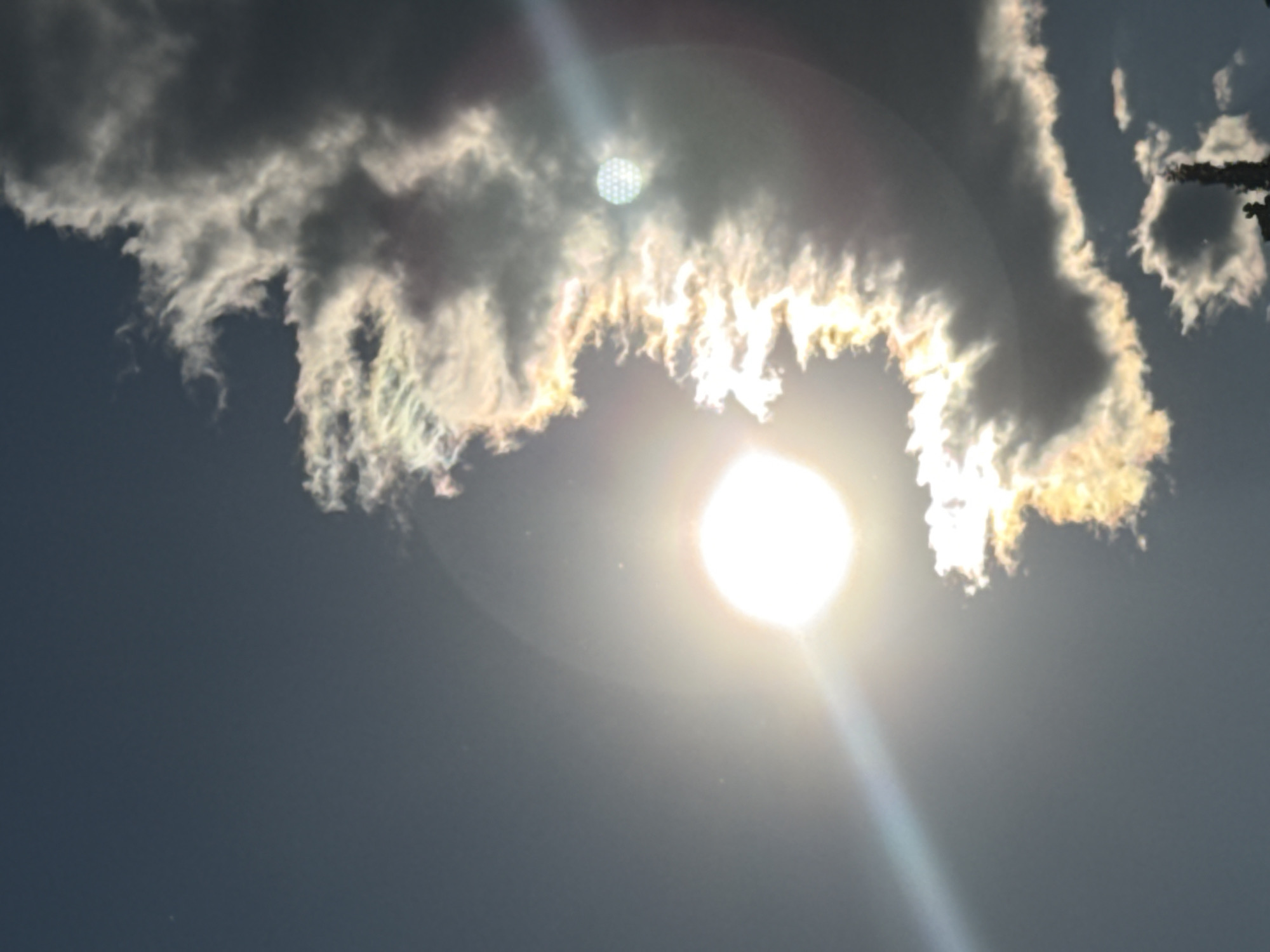 A bright sun is visible in a dark blue sky, with a sun flare extending to the left. A large, dark cloud dominates the right side of the image, with its edges illuminated by the sun, creating a rainbow effect. Smaller clouds are scattered across the sky. A thin, dark object, possibly a tree or structure, is visible at the bottom center of the image, silhouetted against the sky. The image appears to be taken in Stara Zagora, Bulgaria, as indicated by the file name.