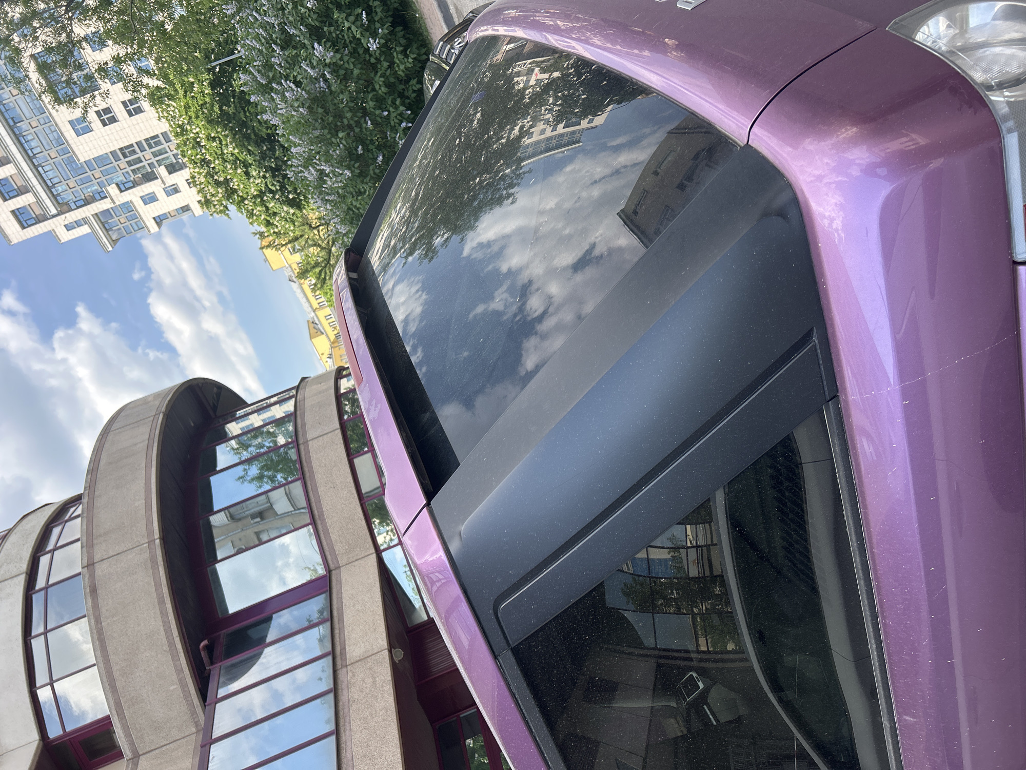 The image shows a close-up of a metallic purple car parked in an urban environment. The car's windshield reflects the surrounding buildings and sky. A modern, rounded building with multiple glass windows is visible to the left. Behind the car and above the building, other structures and foliage are discernible. The sky is partly cloudy. The location appears to be a city with contemporary architecture.