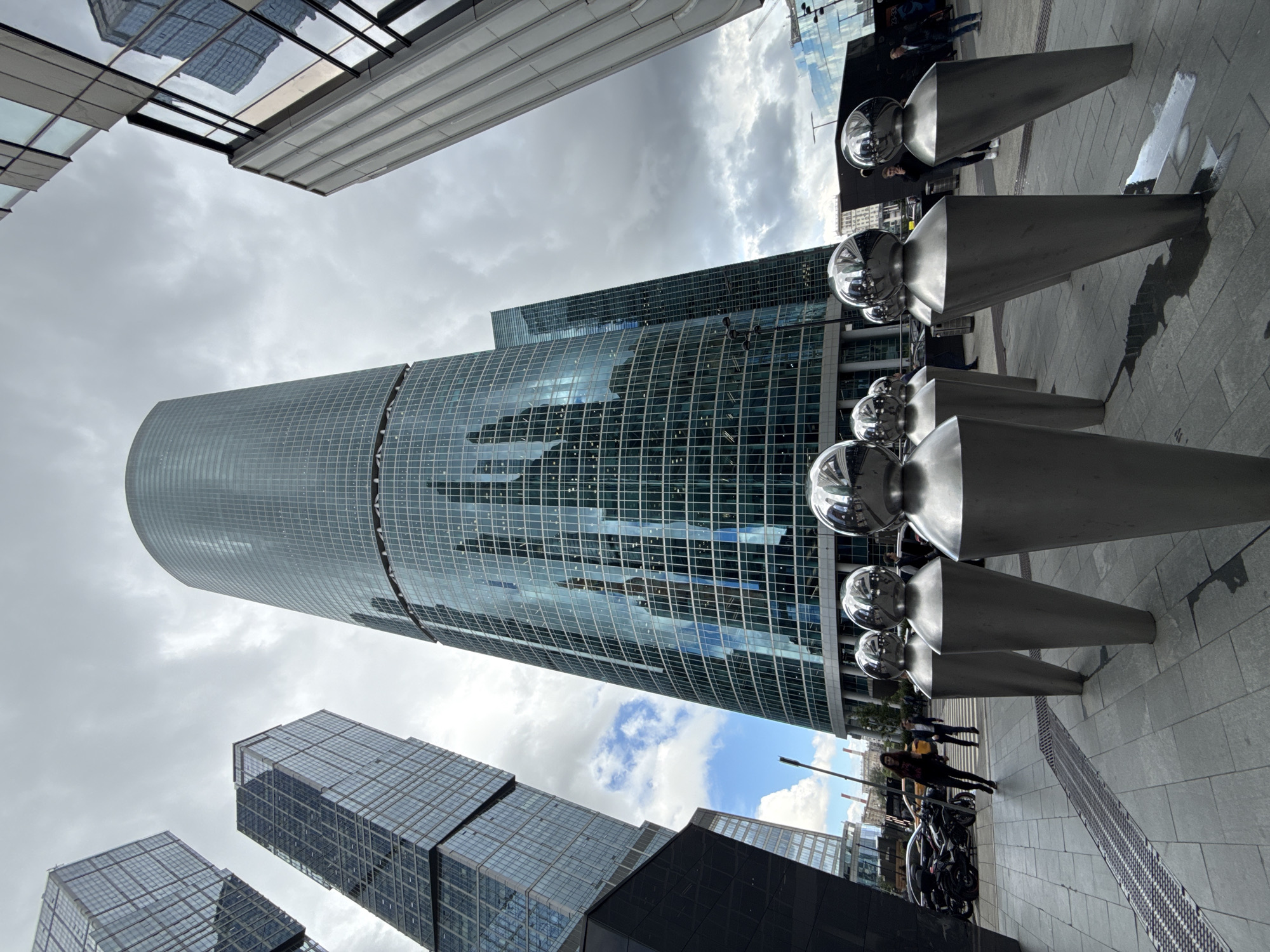 The image depicts the Moscow International Business Center, also known as Moscow City, a business district in Moscow, Russia. In the foreground, several large, metallic, abstract sculptures resembling human figures are arranged on a paved area. The sculptures have conical bases and spherical heads that reflect the surrounding environment. Behind the sculptures, a massive, cylindrical skyscraper dominates the center of the frame. This skyscraper, with its glass facade, reflects the cloudy sky and other buildings. To the left and right of the central skyscraper are other modern skyscrapers with glass exteriors. Several people are visible in the scene, including a woman walking past the sculptures on the left, and a couple of men standing near the sculptures and buildings on the right. Cars and motorcycles are also present in the background. The sky is overcast with some patches of blue sky and clouds, suggesting a mixed weather day. The overall impression is one of a modern, urban landscape characterized by large-scale architecture and public art.