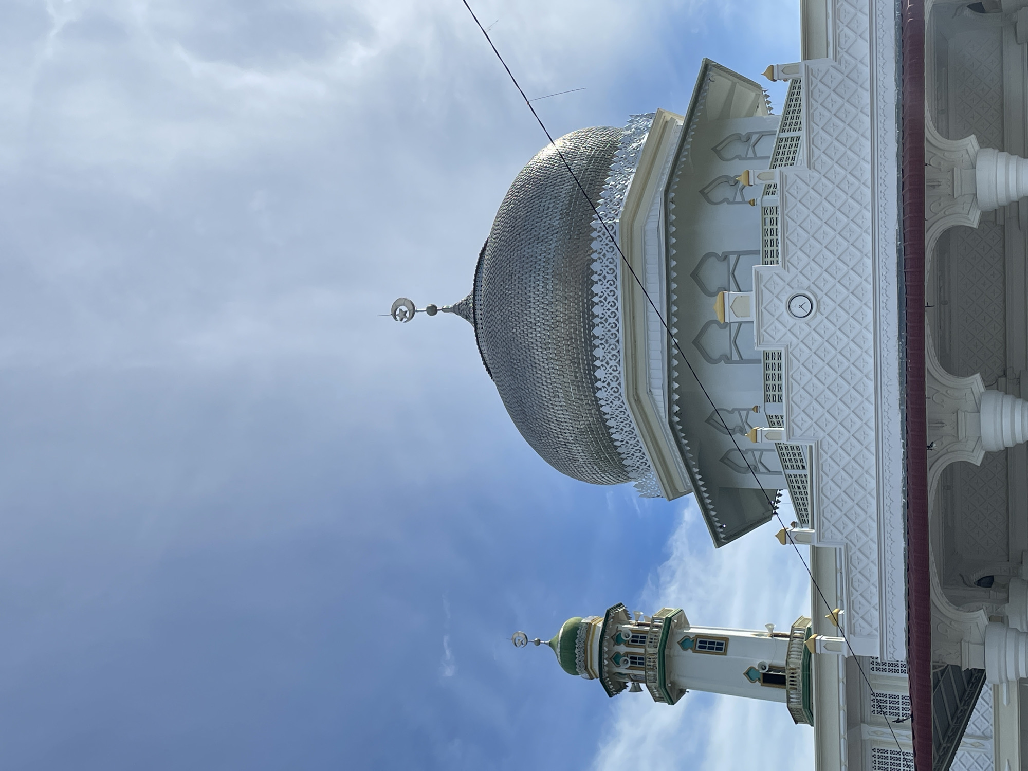 The original image, oriented sideways (rotated 90 degrees clockwise), presents an architectural view of the Mesjid Reubee mosque in Indonesia, captured from a low angle against a bright, partially cloudy blue sky.
The most prominent feature is a large, metallic silver-colored dome on the main mosque building, covered in a detailed, scale-like pattern that reflects the light. The base of the dome is adorned with white, intricate latticework and decorative trim. To the right of the dome (when viewed upright), the mosque's white facade features deeply carved geometric patterns and arched window-like structures with golden accent spires. A small, round clock face is visible on one of the patterned sections of the wall, though its numbers are not discernible.
Further down and to the left of the main dome (when viewed upright), a tall, slender minaret rises towards the sky. The minaret is predominantly white, featuring distinct green and gold-colored sections, multiple balconies with railings, and arched window details. Both the main dome and the minaret are topped with crescent moon and star finials, symbolic elements in Islamic architecture. Thin black lines, possibly power lines or wires, stretch diagonally across the sky in the upper portion of the image. The bright daylight suggests it is daytime, with soft shadows highlighting the architectural details. No people or specific activities are visible in the still scene.