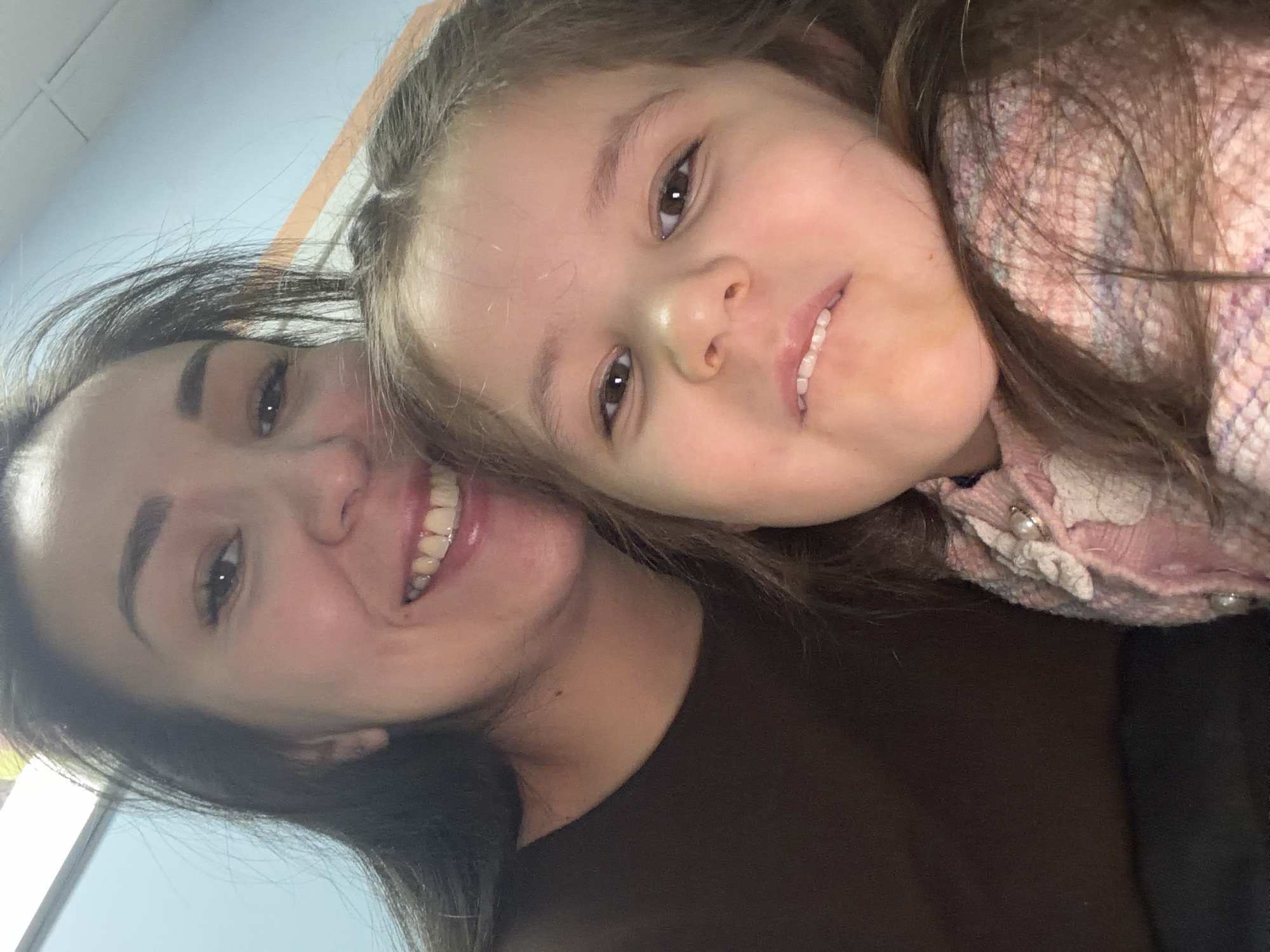 A selfie photograph depicts a woman and a young girl. The woman is smiling broadly with visible teeth. The young girl appears to be resting her head on the woman. The woman is wearing a dark brown top. The girl is wearing a light pink cardigan. The background consists of a light blue wall. There is no indication of the location being Korolyov, Russia.