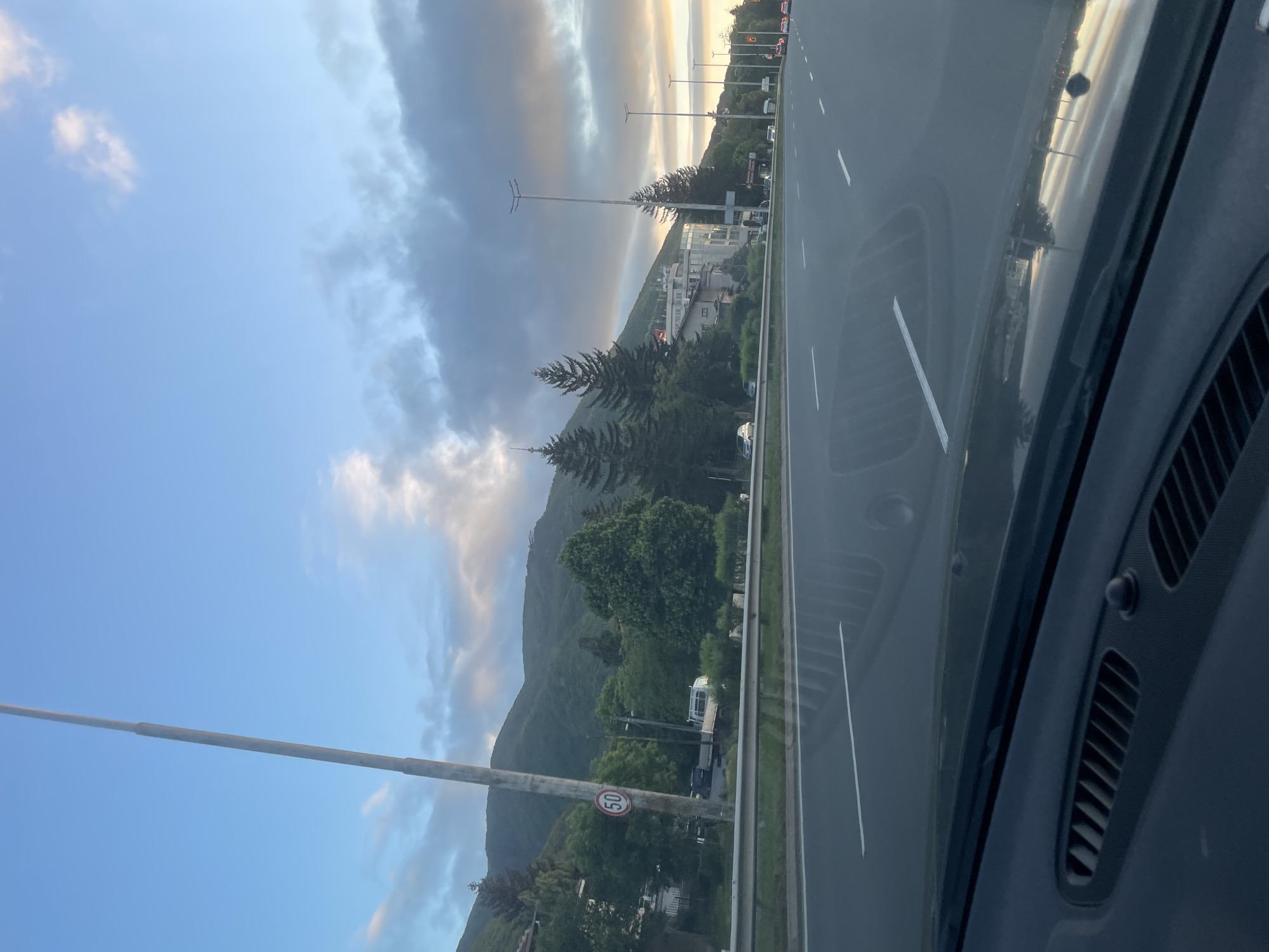 The image shows a view from inside a car on a multi-lane highway. A mountain range dominates the left side of the frame, with a mix of trees and buildings at its base. A street lamp is visible on the left side of the road. A speed limit sign of 50 is visible next to the road. The sky has a gradient of blue and clouds, with a hint of sunrise or sunset on the horizon. The road is smooth and gray. The car's interior, specifically the dashboard and air vents, are visible on the right side of the frame.