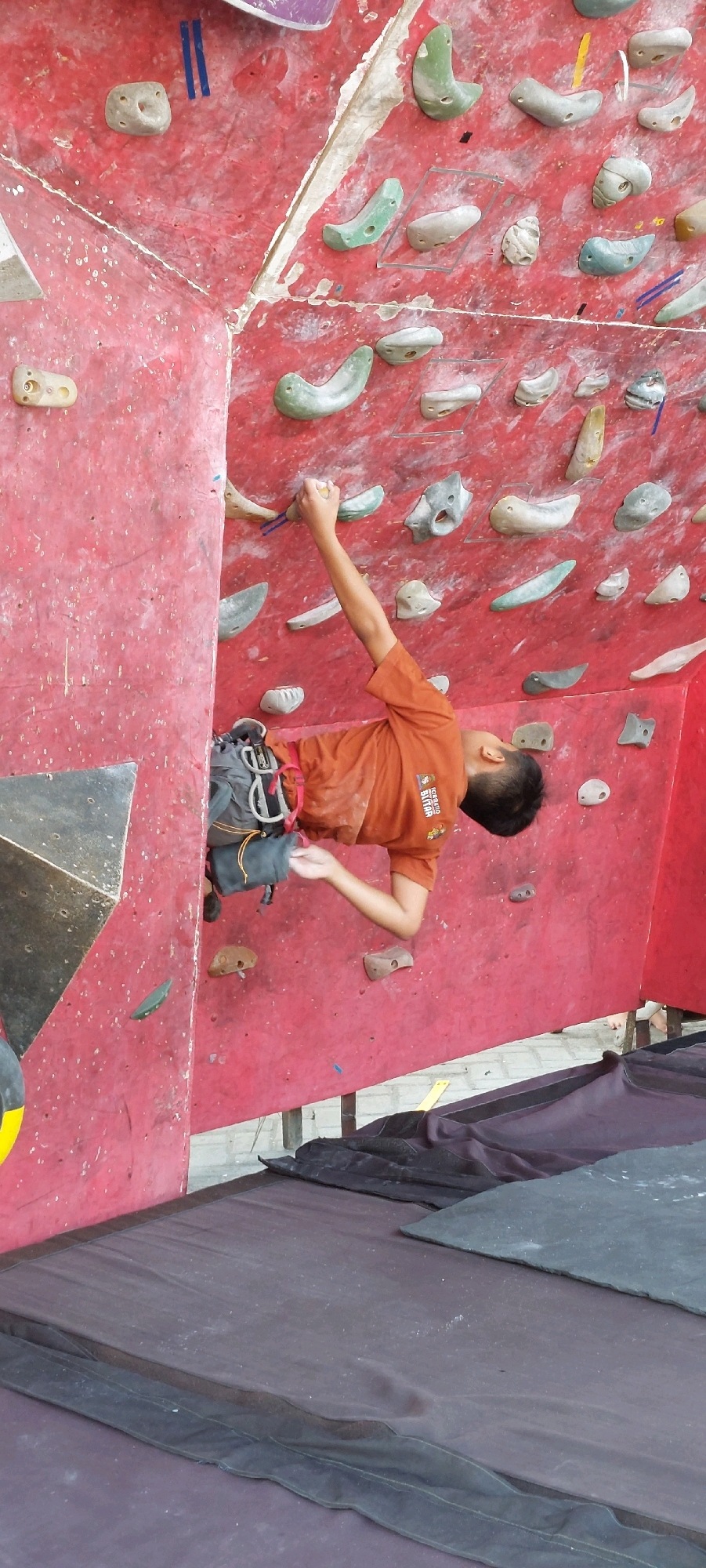 A young person is rock climbing on an indoor climbing wall. They are wearing an orange t-shirt and a climbing harness. Their right arm is extended upwards, gripping a climbing hold. The climbing wall is red and has various colored climbing holds of different shapes and sizes. Below the wall, there is a large padded mat. The scene is viewed from a slightly elevated perspective.