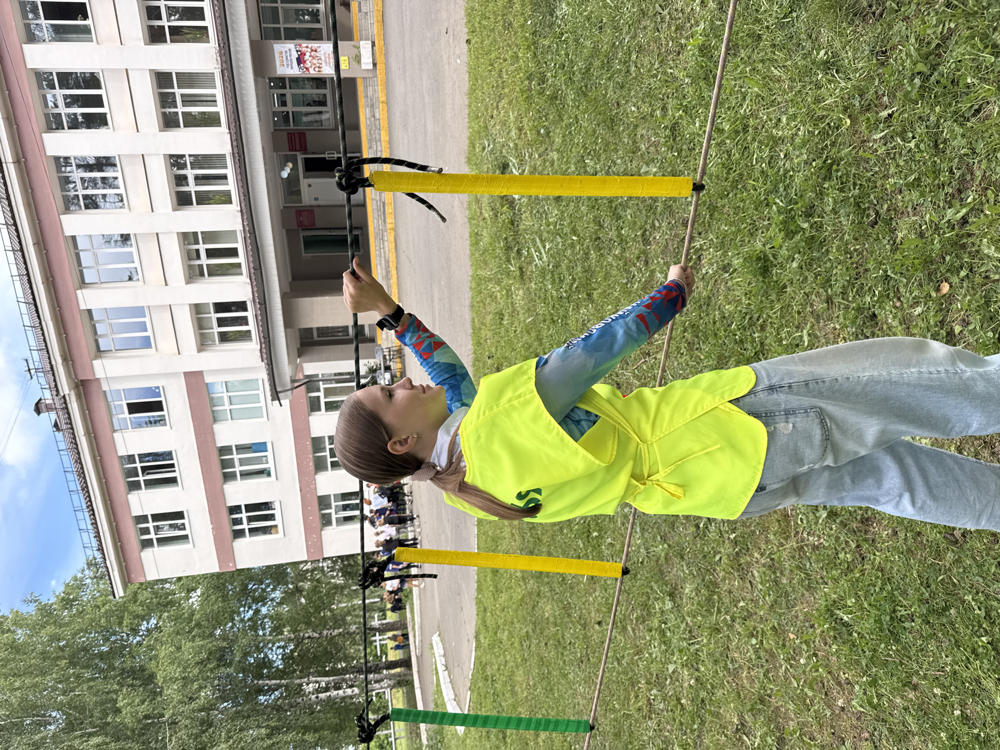 The image depicts a young woman participating in an outdoor activity resembling a rope course, possibly in the city of Chaikovsky, Russia, given the architectural style of the building in the background which is characteristic of Russian educational institutions. The woman is wearing a bright yellow vest over a blue and red patterned long-sleeved shirt and light blue jeans. She is holding onto a rope with both hands, balancing herself, as if walking on a slackline. The rope is supported by two vertical poles, one green and one yellow, wrapped in tape. In the background, a multi-story building with a pink-trimmed roof and numerous windows stands. Other individuals, likely students or participants, are visible in the distance near the building. The ground is covered with green grass, and the sky is partly cloudy. The scene suggests a recreational or educational event taking place on a school campus or a park.
