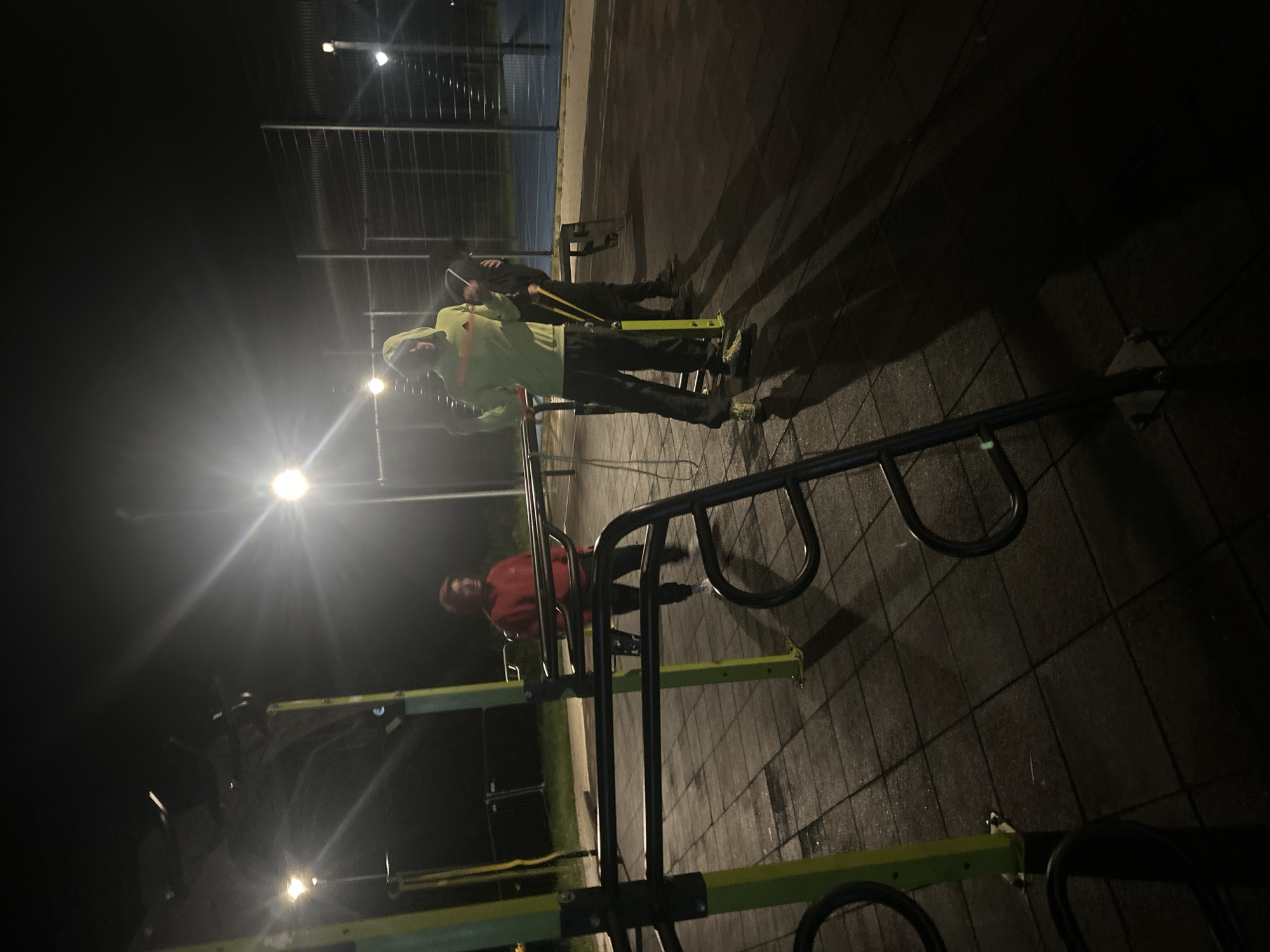 The image depicts a street workout area at night, illuminated by overhead lights. Three individuals are present, with two appearing to be exercising using the equipment. One person wears a light green hooded jacket. The other person further back wears dark clothing. The scene includes multiple pieces of outdoor exercise equipment with yellow and black support structures. The ground is covered in dark paving tiles. In the background, a fence separates the workout area from an undefined space, where a structure with lights is visible. The exact location within Burgas, Bulgaria, cannot be determined from the given image alone.