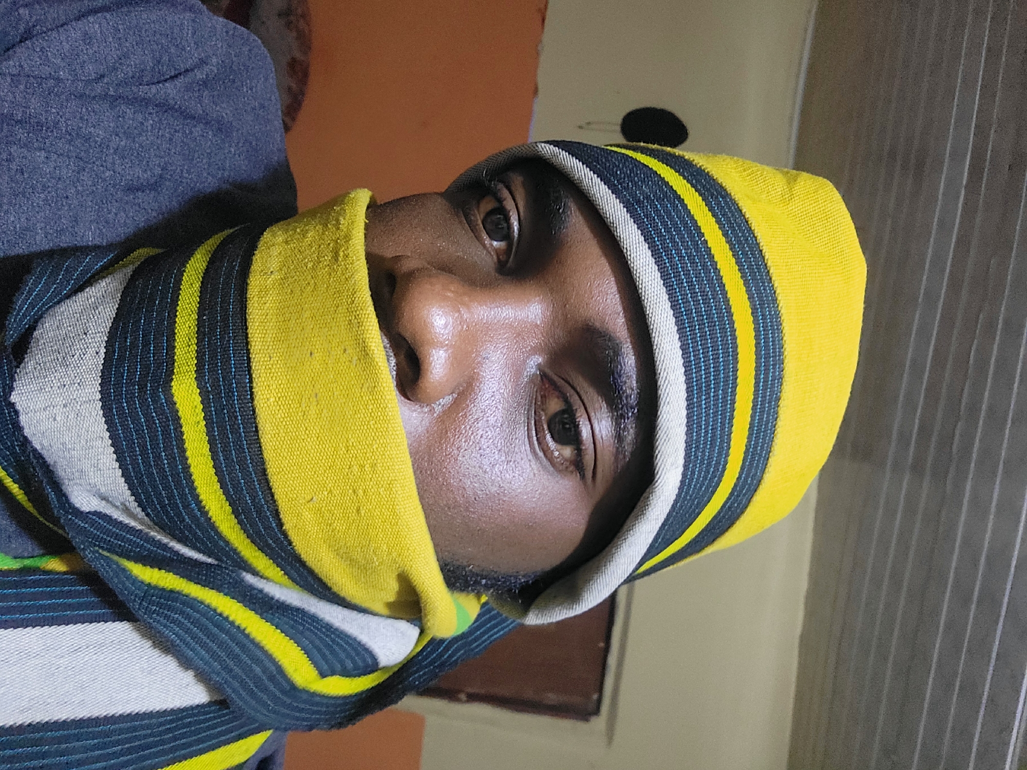 A close-up, waist-up selfie shows a person wearing a yellow, blue, and gray striped beanie and scarf that covers their lower face. The person's eyes are visible, and they appear to be looking directly at the camera. The background is blurry and dimly lit, with a textured ceiling and an orange wall. A dark, circular object is visible on the wall behind the person's head, possibly a light fixture. The scene appears to be indoors.