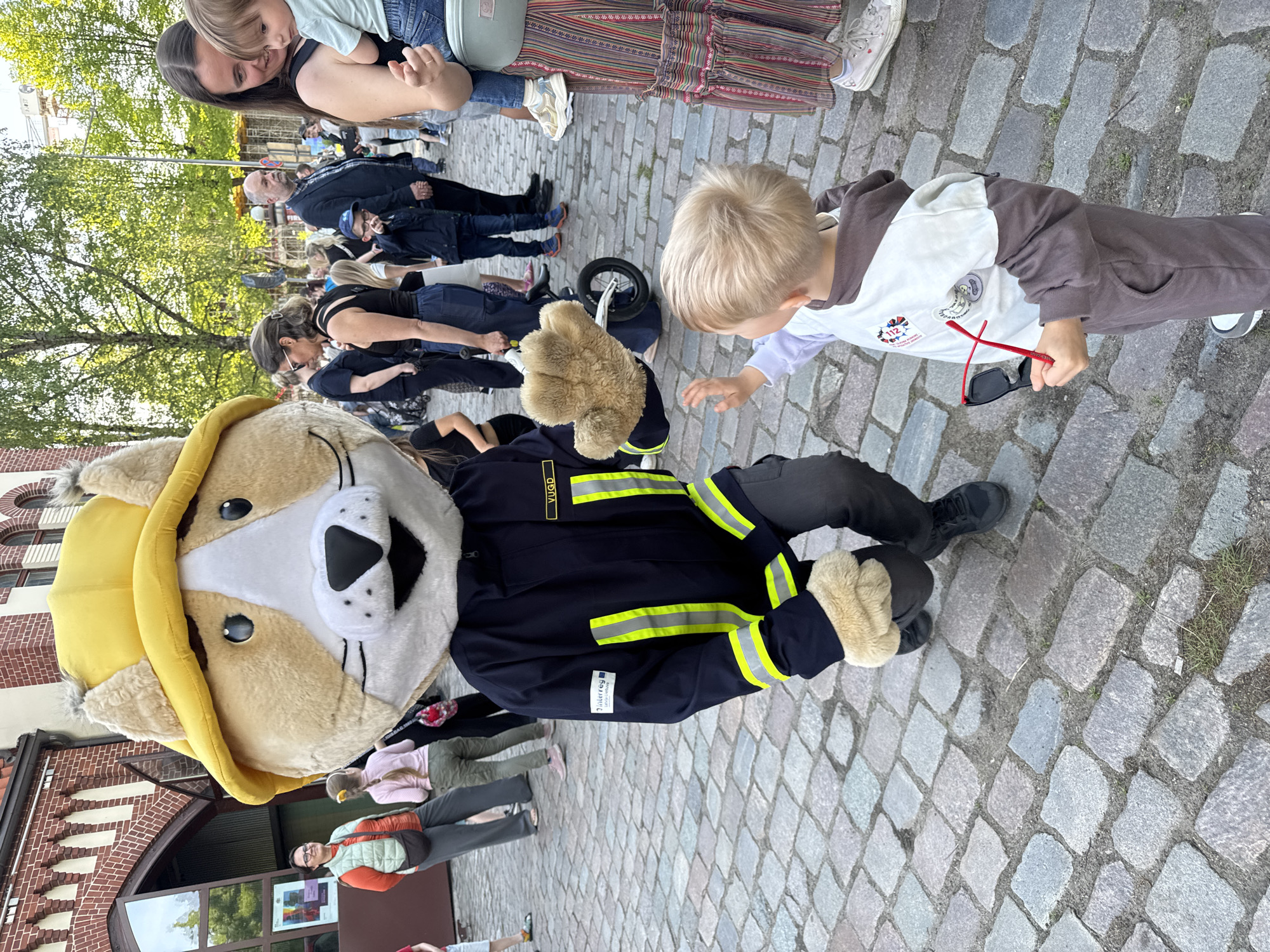 A vertical, medium shot shows a group of people and a mascot on a cobblestone surface.
A young child with blonde hair, wearing a brown and white hooded sweatshirt and grey sweatpants, holds a pair of red and black sunglasses and reaches towards a large, stuffed mascot. The mascot, representing a canine figure with a yellow fire helmet and dressed in a black firefighter's uniform with yellow reflective stripes, is seated on the cobblestone. A crowd of people is in the background, some standing and some seated. The cobblestone surface dominates the foreground.
A brick building with arched windows and a glass entryway is in the background on the left. The city of Riga, Latvia is confirmed by the 'YUUGD' lettering on the uniform of the mascot. The scene captures an interaction between a child and a costumed character in a public space.