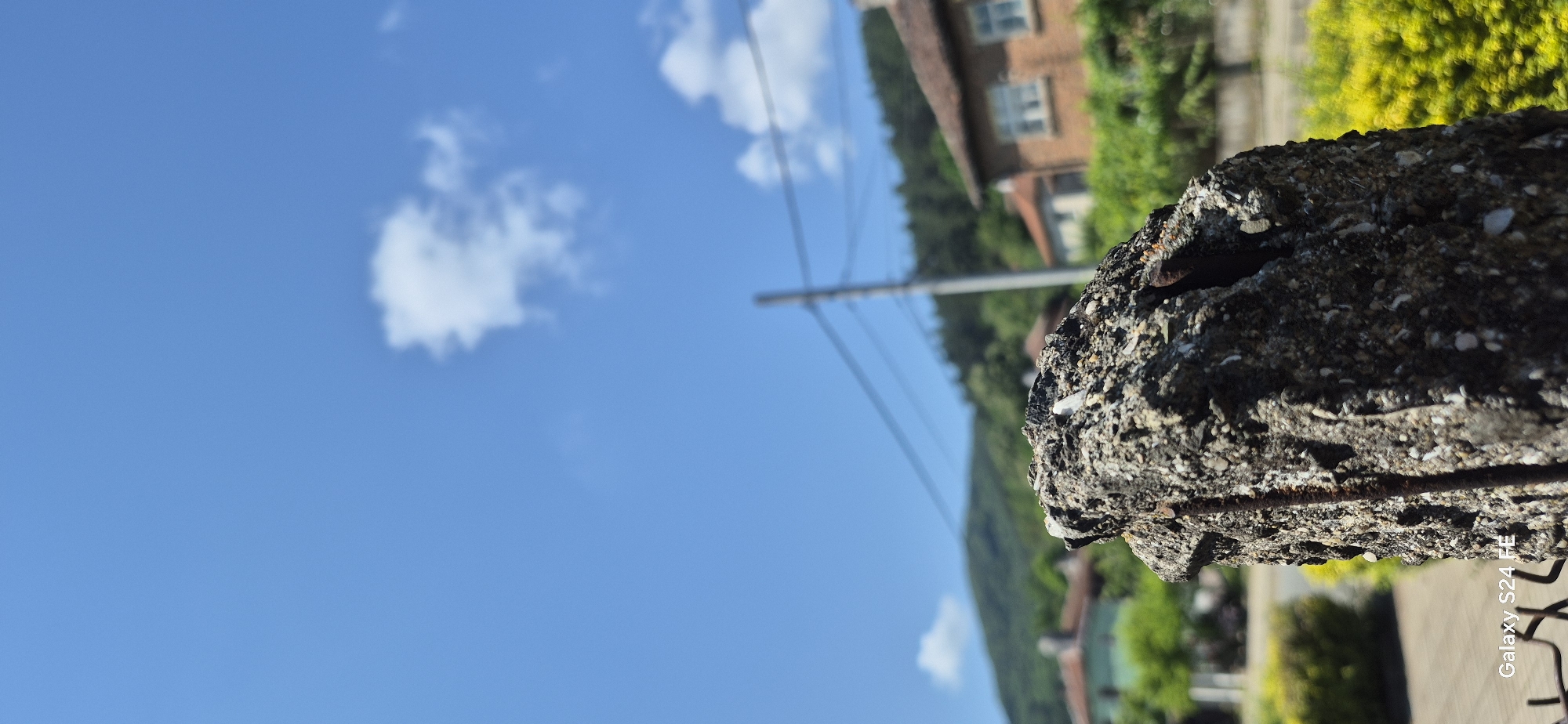 The image is a close-up shot of a weathered concrete post, possibly a fence post, against a backdrop of a clear blue sky with a few wispy clouds. The post is rough and textured, with visible aggregate and a rusty metal rebar protruding from its side. In the mid-ground, a residential area is visible, with houses featuring terracotta roofs, lush green trees, and a winding road. Beyond the houses, rolling green hills covered in dense forest rise up, creating a scenic mountainous landscape. The overall impression is of a peaceful, rural setting in a town like Malko Tarnovo, Bulgaria, where nature and human habitation coexist. There are no people or animals visible in the frame, and no specific actions are taking place. The image appears to be taken during daylight hours, with bright sunlight illuminating the scene.