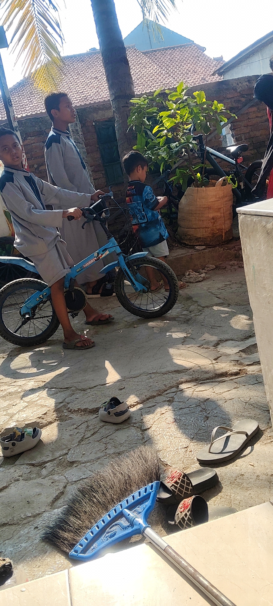 In Mekarmukti, Indonesia, a young boy in a gray tunic and shorts rides a blue bicycle with "THOMAS" written on the frame. Another boy in a blue t-shirt with a wave pattern and white shorts stands beside the bicycle. A third boy, also in a gray tunic and shorts, stands behind the boy on the bicycle. They are on a paved ground in what appears to be a residential area with brick walls and tiled roofs visible in the background. A palm tree is to the left of the frame, and green foliage from a potted plant is to the right. Scattered on the ground are several pairs of footwear, including flip-flops, slide sandals, and clogs. A broom with a blue plastic head and metal handle lies in the foreground. The scene is illuminated by bright sunlight, casting shadows on the ground.