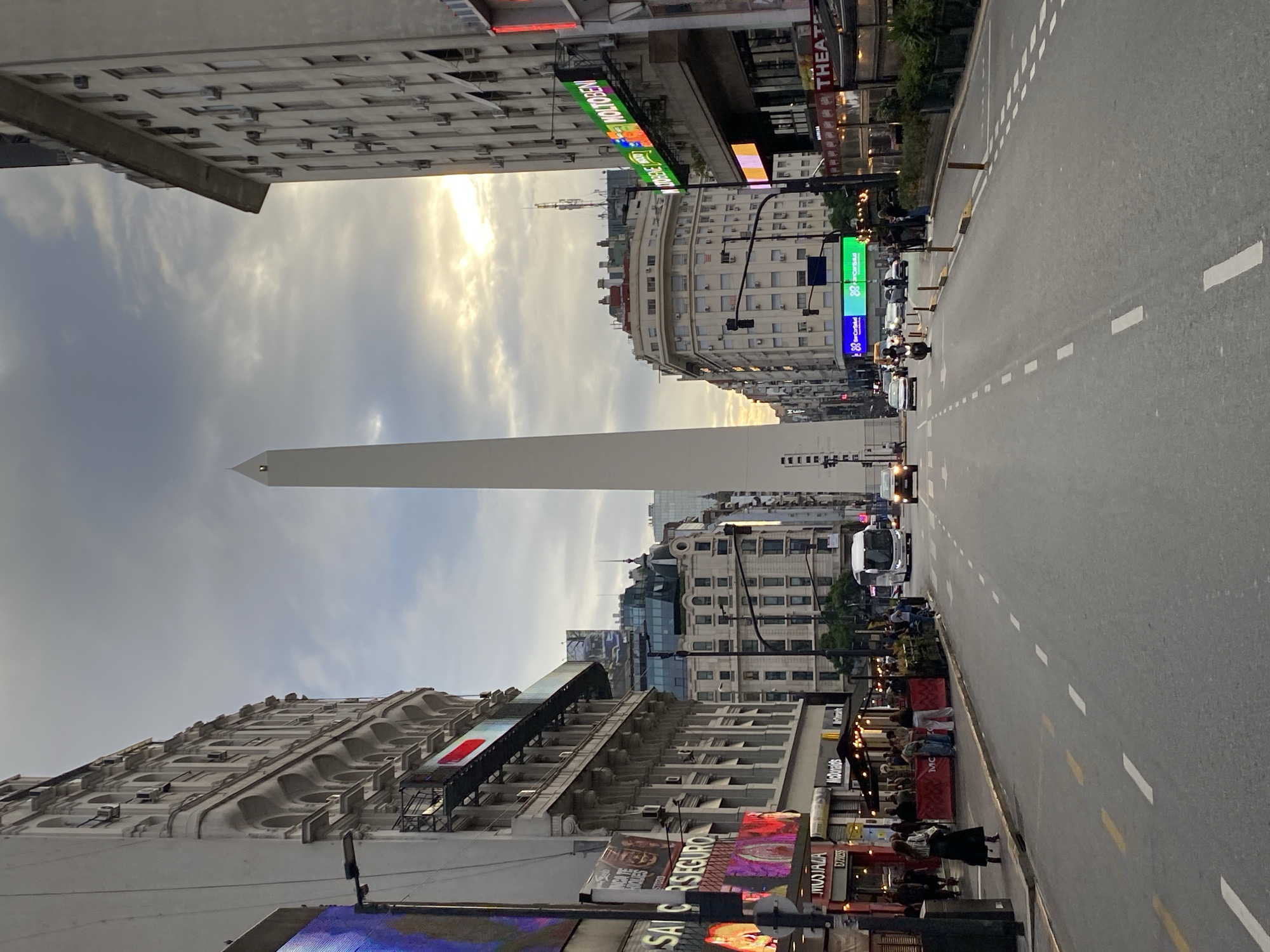 The image depicts a street scene in Buenos Aires, Argentina, specifically looking southeast down Avenida Corrientes towards the Obelisco.
In the mid-ground, the white, monolithic Obelisco de Buenos Aires stands centrally, its apex silhouetted against a partly cloudy sky. The sky above is overcast with a bright, warm light source, likely the setting sun, visible behind the obelisk and to its right.
The foreground is dominated by a multi-lane asphalt road with white dashed lane markings. Several vehicles are present: a white bus, a black car, a motorcycle, and other passenger vehicles, primarily in the right lanes, moving away from the viewer.
On the left side of the street, a multi-story building features classical architecture with arched window frames and decorative elements. Its lower levels host commercial establishments, including visible signage for "SAI SEGURO," "MUSTAFA EXPRESS," and a McDonald's restaurant with an outdoor seating area enclosed by red barriers. Several pedestrians are visible on the sidewalk and within the outdoor dining space. A large, illuminated digital billboard is partially visible on the far left.
On the right side of the street, another multi-story building with a more utilitarian design displays numerous external air conditioning units. Commercial signs, including a green "MOLTOBENI" sign and a partial "THEATER" sign, are mounted on its facade. Pedestrians occupy the sidewalk, and outdoor seating areas are also present. A black traffic light signal is visible on the right.
Further down the street, additional buildings line both sides, varying in height and architectural style, some topped with antenna arrays. The overall scene suggests a busy urban environment during late afternoon or early evening.