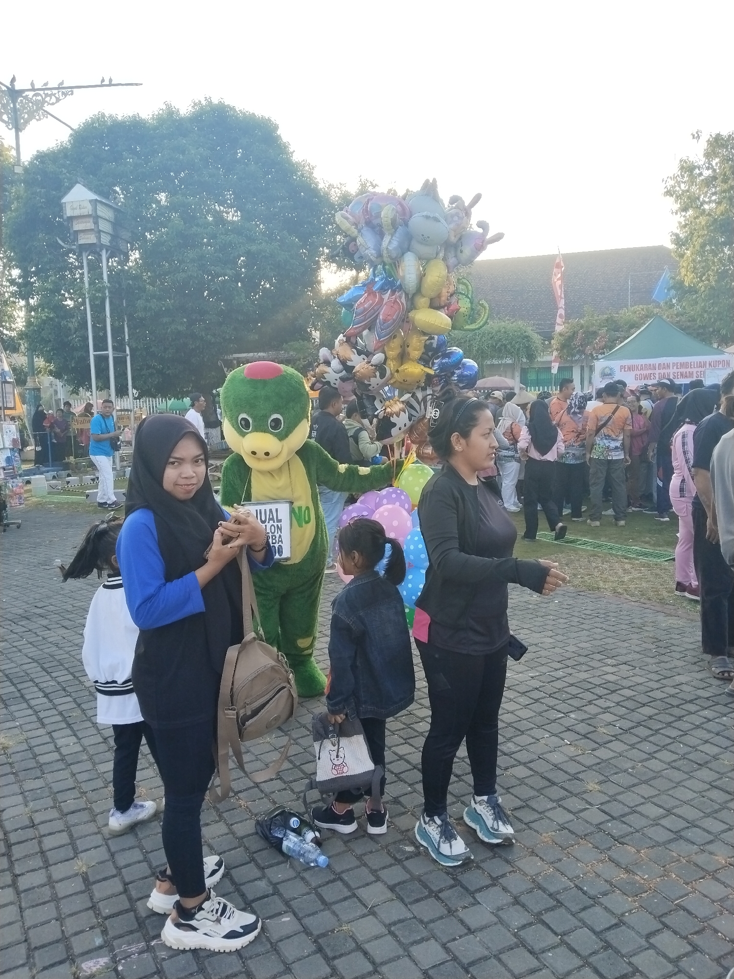 This image captures an outdoor event in Sragen, Indonesia, with multiple people, a mascot, and various stalls. In the immediate foreground, the ground is paved with rectangular grey interlocking pavers, with small patches of grass visible in the cracks. A clear plastic water bottle and a dark object, possibly a small bag or a piece of fabric, rest on the ground near the left side. On the left, a woman wearing a black hijab, a royal blue long-sleeved shirt, and black leggings stands facing slightly towards the camera. She carries a tan canvas crossbody bag and holds a small, dark item (possibly a phone) in her hands, looking at the camera with a subtle smile. She wears chunky white, black, and tan athletic shoes. Partially obscured behind her is a young girl with dark hair in pigtails, wearing a white long-sleeved shirt with black trim and dark pants, along with light-colored sneakers. Central to the midground is a person in a full-body green mascot costume resembling a cartoon bird or turtle, featuring a red cap, yellow beak, and large white eyes. The mascot holds a white sign that reads "JUAL BALON RP BA 00" (Sell Balloons RP BA 00). To the right of the mascot, another woman in a black long-sleeved top, black leggings, and white and blue athletic shoes is captured in a gesturing pose, with her arms slightly outstretched and palms open, facing towards the right side of the frame. A hint of a pink garment is visible beneath her black top on her left hip. To her left, a second young girl, also with dark hair in pigtails, wears a blue denim jacket over dark clothing and carries a small white tote bag with a cartoon character design. A large cluster of Mylar balloons, featuring various animal shapes (such as a dinosaur, a rabbit, and a bear) and other characters in multiple colors (silver, gold, blue, pink, green), rises behind the mascot and the central figures. A smaller grouping of pink and blue polka-dotted balloons is near the feet of the gesturing woman. In the background, numerous people are visible, some walking and others standing. On the far right, a white tent with a green roof displays partially visible Indonesian text on its side: "PENUKARAN DAN PEMBELIAN KUPON GOWES DAN SENAM SE..." (Coupon Exchange and Purchase for Cycling and Aerobics...). A red and white striped flag or