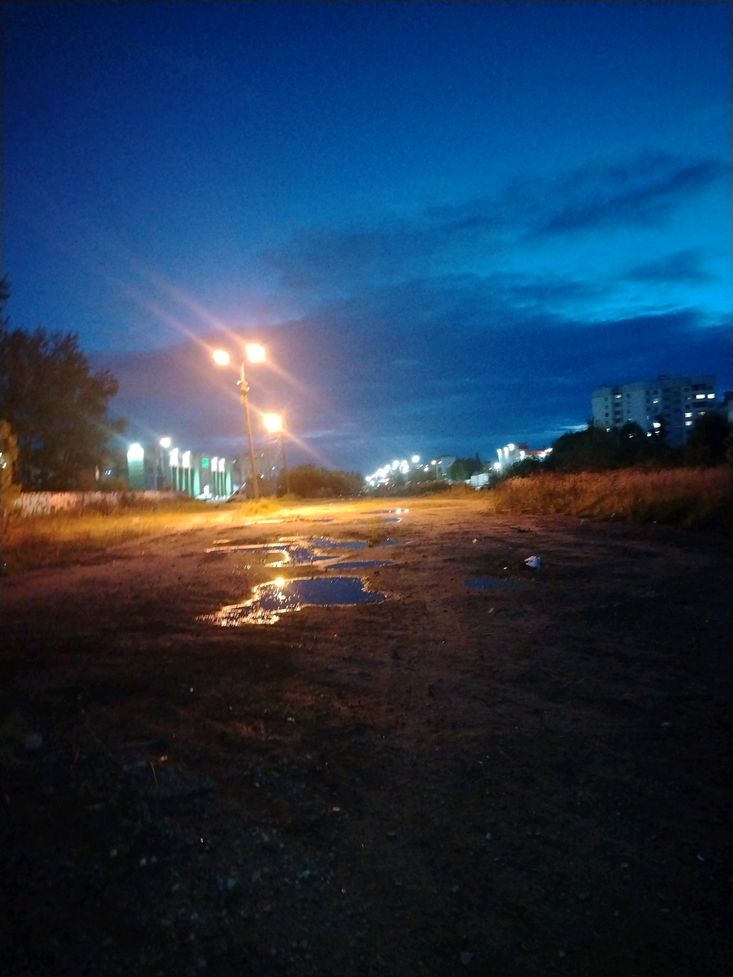 The image depicts a view of a dirt road with puddles reflecting the light of street lamps. The sky is a deep blue, characteristic of twilight, with some clouds visible. In the background, there are buildings, including what appears to be a multi-story apartment building, and other structures with illuminated windows, suggesting an urban or suburban environment. Streetlights are a prominent feature, casting bright light and creating lens flares. There are trees and dry vegetation on the sides of the road. The overall scene suggests an outdoor location at dusk or night. Based on the visual cues, the location is identified as being on the outskirts of the city of Yaroslavl, Russia, specifically near the intersection of Moskovsky Prospekt and Chernosolskoy Lane. The presence of common architectural styles and vegetation further supports this identification. There are no visible people or animals in the scene. The ground is uneven and appears to be unpaved, with evidence of recent rainfall due to the puddles.