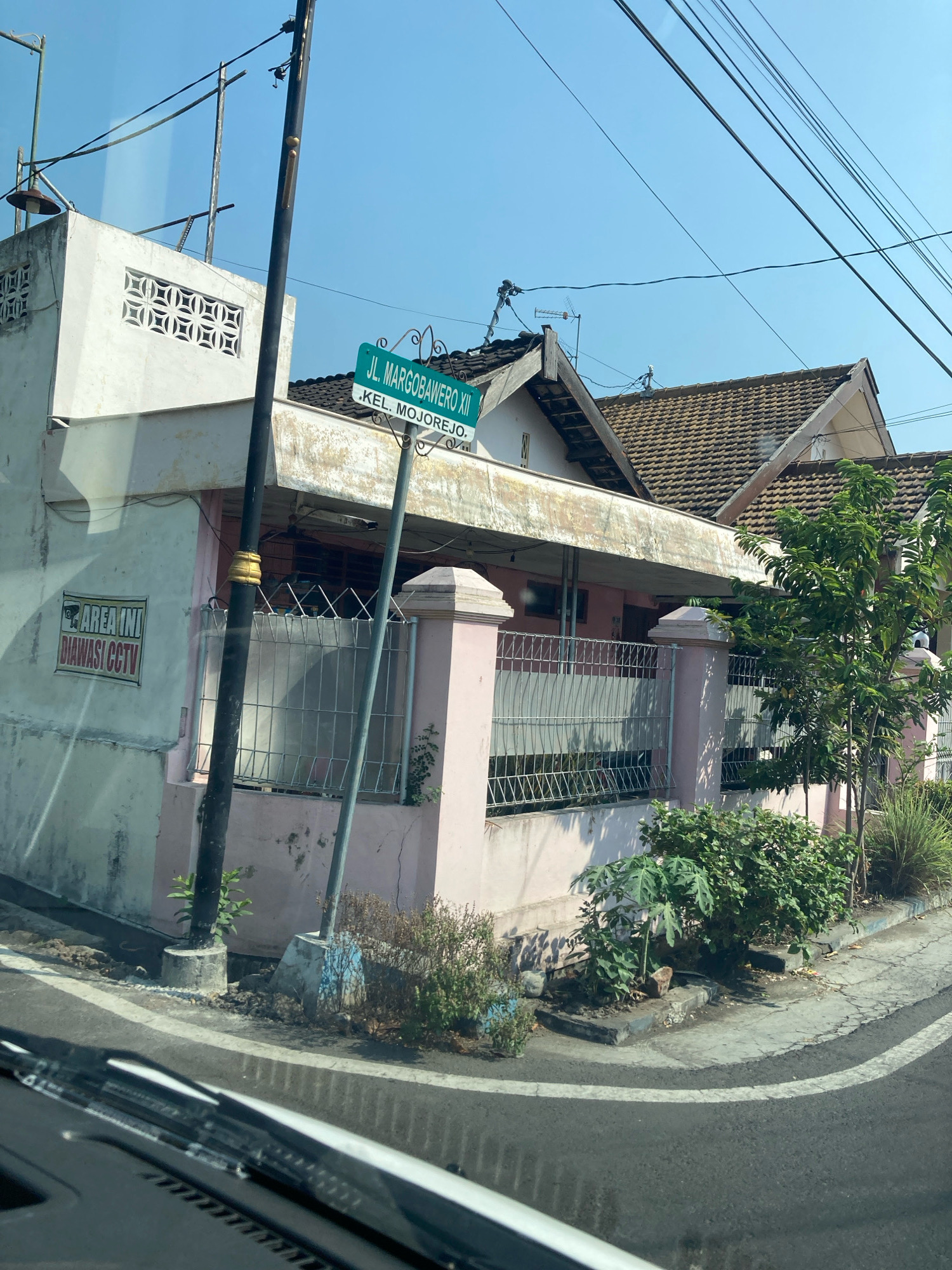 The image shows a street view in an urban environment. The foreground is dominated by the dashboard and windshield of a vehicle, suggesting the photo was taken from inside a car. The middle ground features a light pink house with a white facade and a tiled roof. A street sign in green reads "JL. MARGOBAWEROK XII KEL. MOJOREJO". A prominent pole stands in front of the house, with several electrical wires running overhead. To the left of the house, a white sign with red text states "AREA INI DIAWASI CCTV". Various plants and bushes are visible in front of the house, bordering the street. The sky is clear and blue, indicating a sunny day. The road in the foreground is asphalt with white lane markings. The overall scene depicts a residential street.