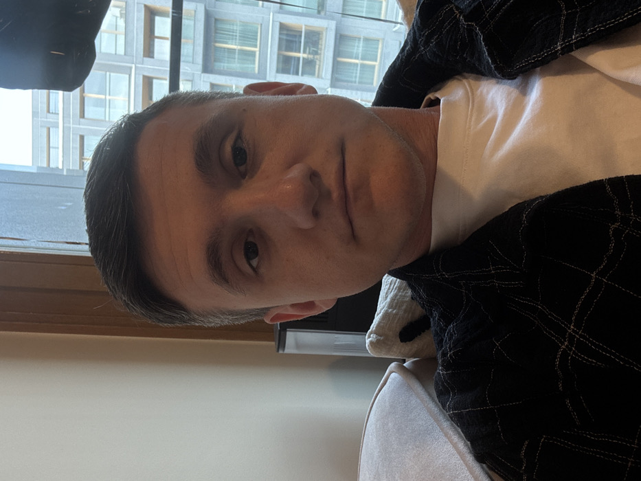A male subject, appearing to be in his 20s or 30s, is centered in a close-up, chest-up indoor portrait. He has short, dark hair, light skin, and a neutral expression, looking directly at the camera. He is wearing a dark, possibly black, plaid or checkered shirt over a white crew-neck t-shirt. To the subject's left, part of a light-colored cushioned surface is visible, suggesting he is seated on a sofa or chair. Behind his left shoulder, a dark, vertical electronic device with a metallic top portion is partially visible. The background features a large window, through which a modern, multi-story building with a grey facade, numerous rectangular windows, and recessed balconies is visible. The external environment suggests an urban setting under an overcast sky. This scene is located in the city of Warsaw, Poland.