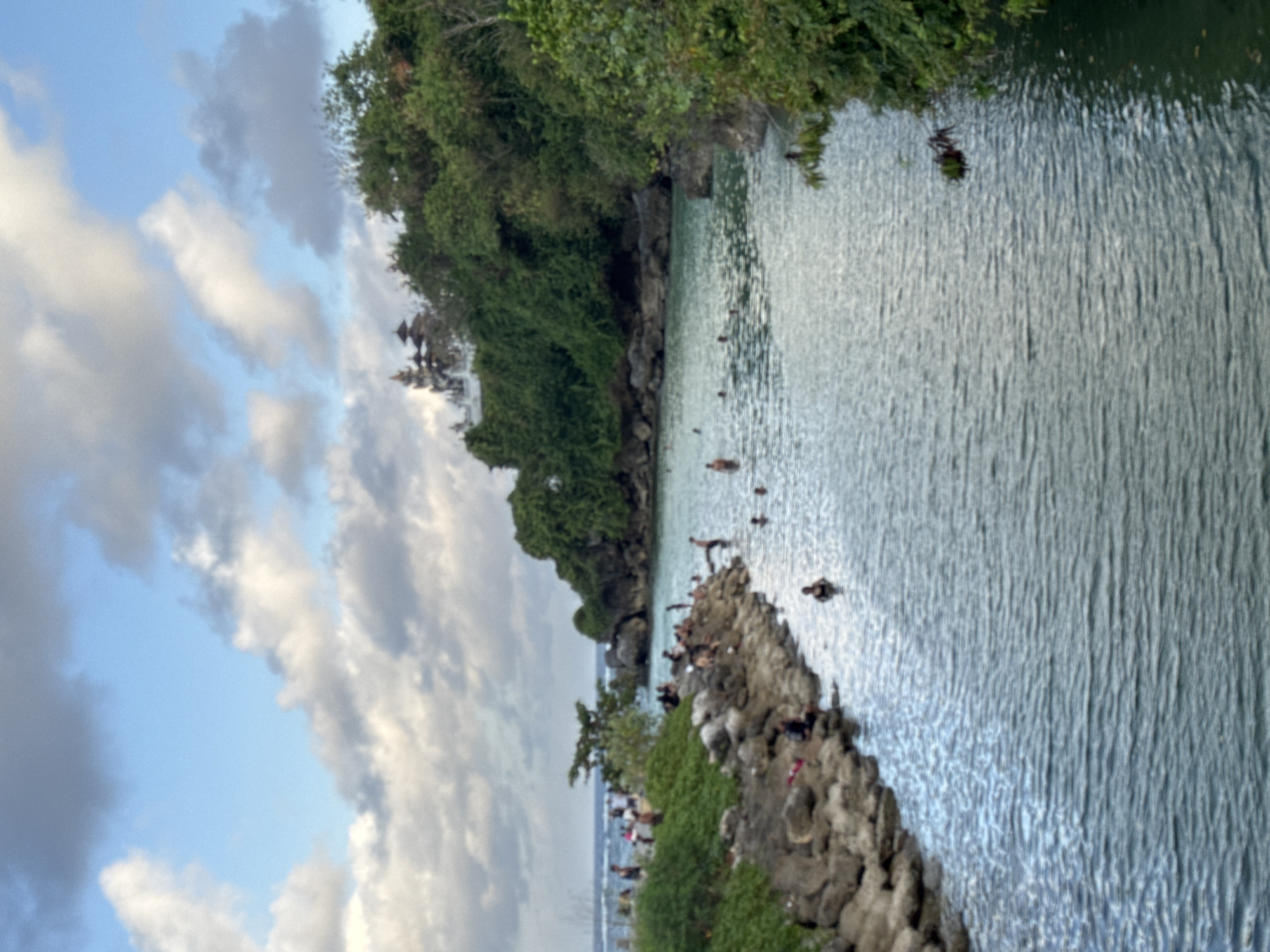 The image shows a coastal scene in Kuta Selatan, Indonesia. The view is of a body of water bordered by lush greenery and rocky terrain. Several people are visible in the water, suggesting swimming or bathing activities. The sky is partly cloudy. The overall atmosphere is tranquil, suggesting a popular tourist or recreational spot. The image is oriented sideways.