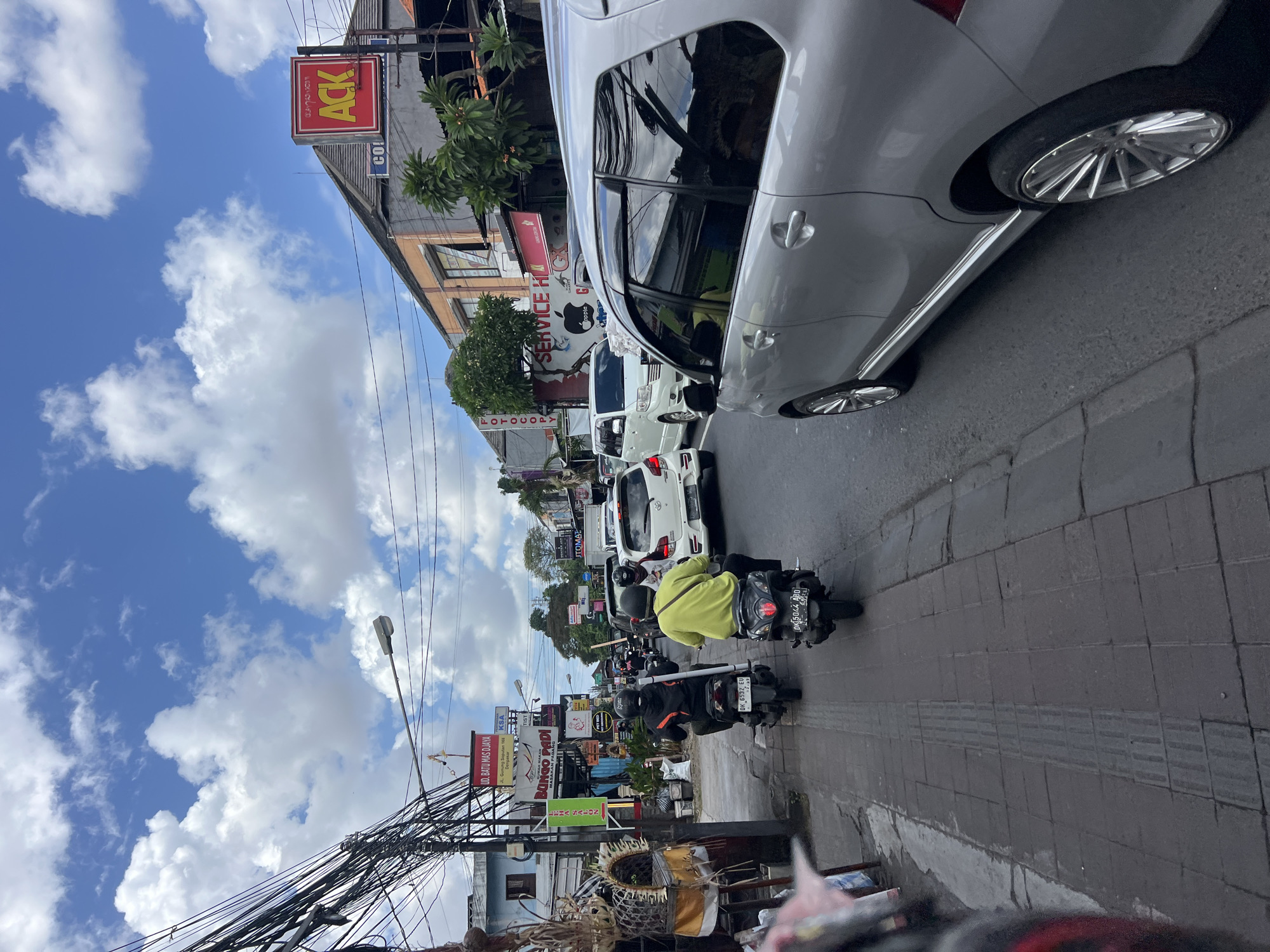 The image depicts a street scene in Denpasar, Indonesia, likely during daylight hours, with a bright sky featuring scattered white clouds. The perspective is from behind a silver car, looking forward down a busy street.
On the left side of the road, a sidewalk is visible, paved with square tiles and featuring tactile paving strips for the visually impaired. Along the sidewalk, there are several shops and businesses with various signs. One sign, in red and yellow, reads "ACK". Another, in green, says "FOTOCOPY". A red sign with white lettering says "BINGO SADI". There are also numerous utility poles with a dense network of electrical wires overhead.
The road itself is paved with asphalt and is moderately congested with traffic. Several motorcycles are prominent. In the foreground, two people are riding motorcycles in the same direction as the camera. The rider in front wears a dark jacket and a helmet, and the rider behind wears a bright yellow jacket and a helmet. Both motorcycles have visible license plates. Further down the road, more motorcycles and cars are visible, including a white car and a white minivan.
Buildings line the right side of the street, displaying a mix of architectural styles and colors. These buildings appear to house businesses, indicated by signage. One sign has the "Apple" logo and text that reads "SERVICE HI". Another sign, a red rectangle with yellow lettering, is partially obscured but appears to be part of the "ACK" sign mentioned earlier. Trees and other greenery are present, adding some natural elements to the urban landscape. The overall impression is of a typical commercial street in a bustling Indonesian city.