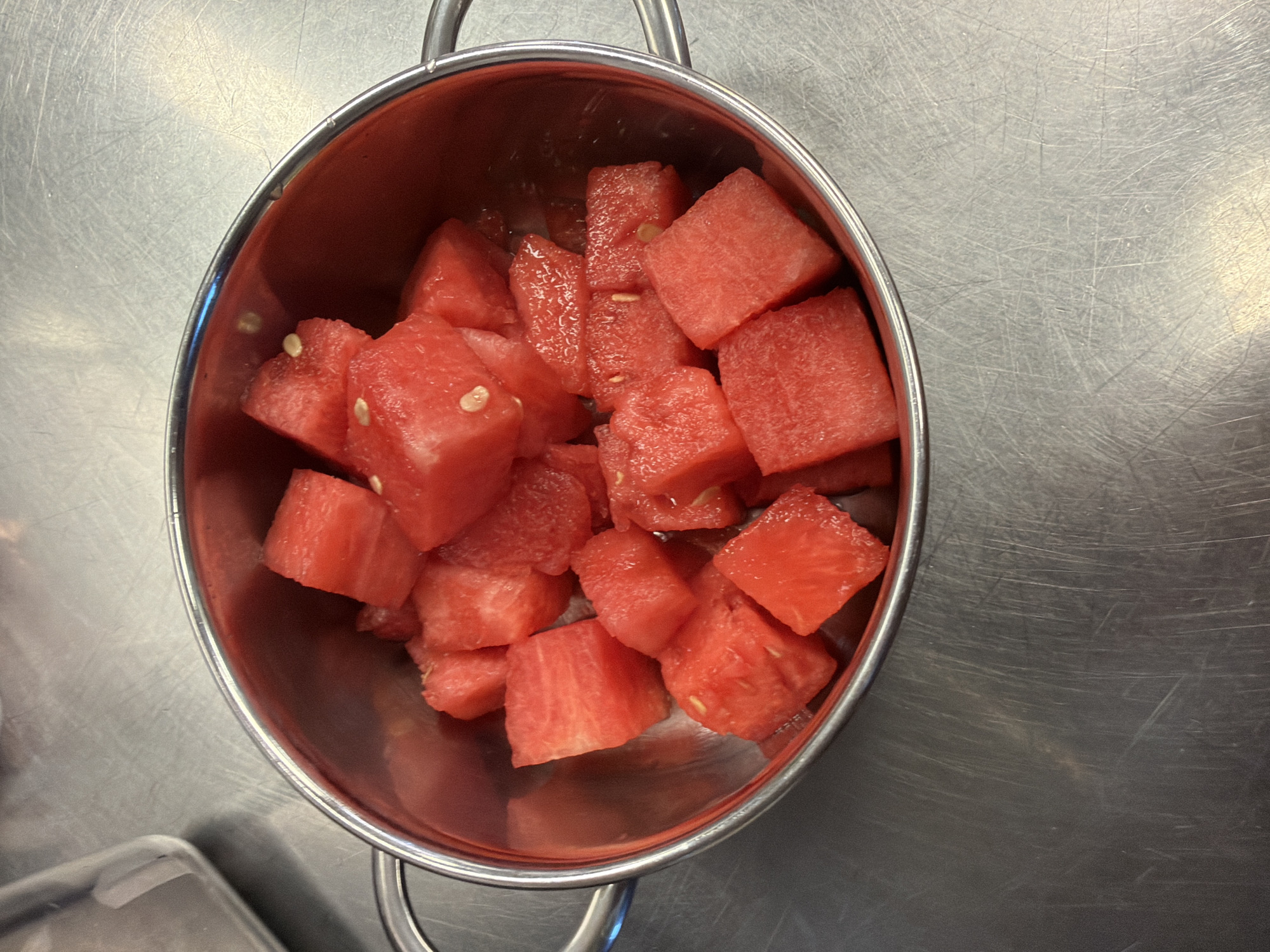 A close-up, top-down view shows a metallic bowl filled with diced watermelon. The watermelon pieces are bright red and contain visible seeds. The bowl has two handles and rests on a brushed metal surface. The background is out of focus and appears to be a kitchen or food preparation area. There are no people or animals visible in the image. The scene is static and appears to be a still life. The location in Rust, Germany, is not visually identifiable from the image.