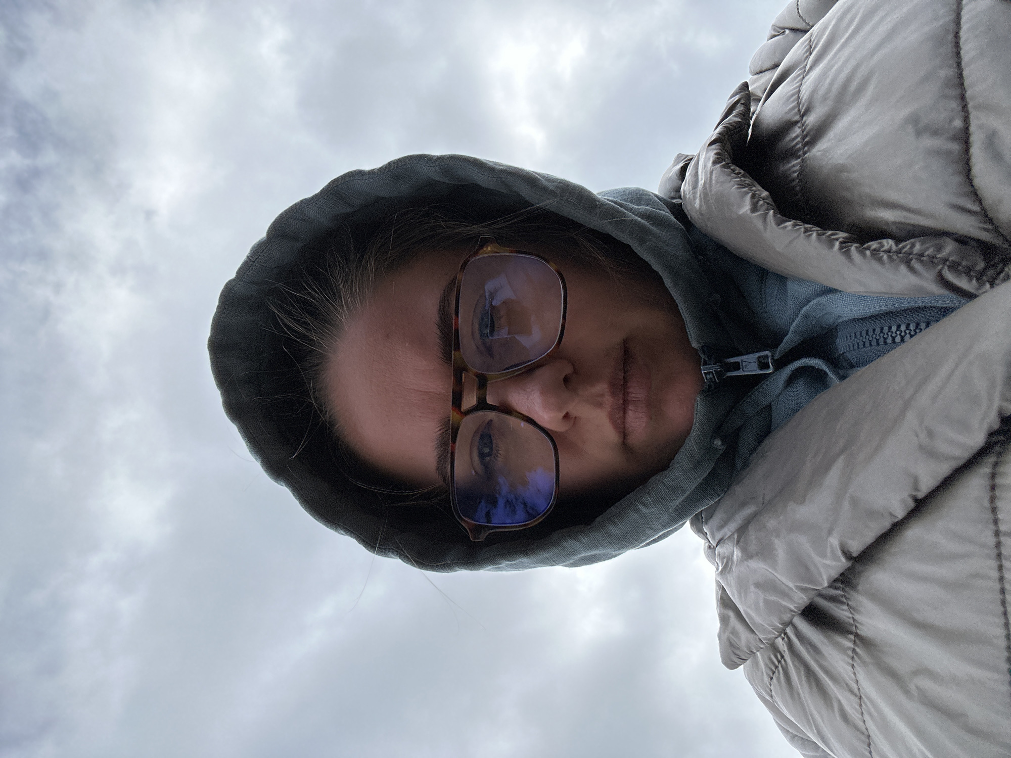 A woman wearing glasses and a hooded jacket is seen from a low angle against a cloudy sky. The location cannot be determined from the image.