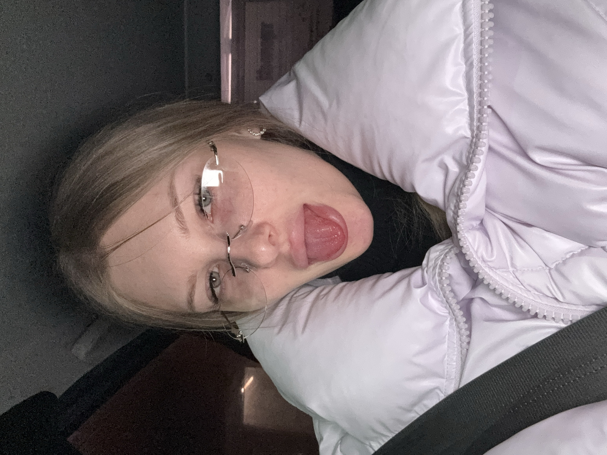 A blonde female, wearing frameless glasses and a lavender puffy coat, is sticking her tongue out at the camera. She is wearing a black turtleneck underneath the coat. A gray seatbelt is visible diagonally across the bottom of the frame. The location is likely inside a vehicle based on the seatbelt and is possibly in Perm, Russia (per provided information).