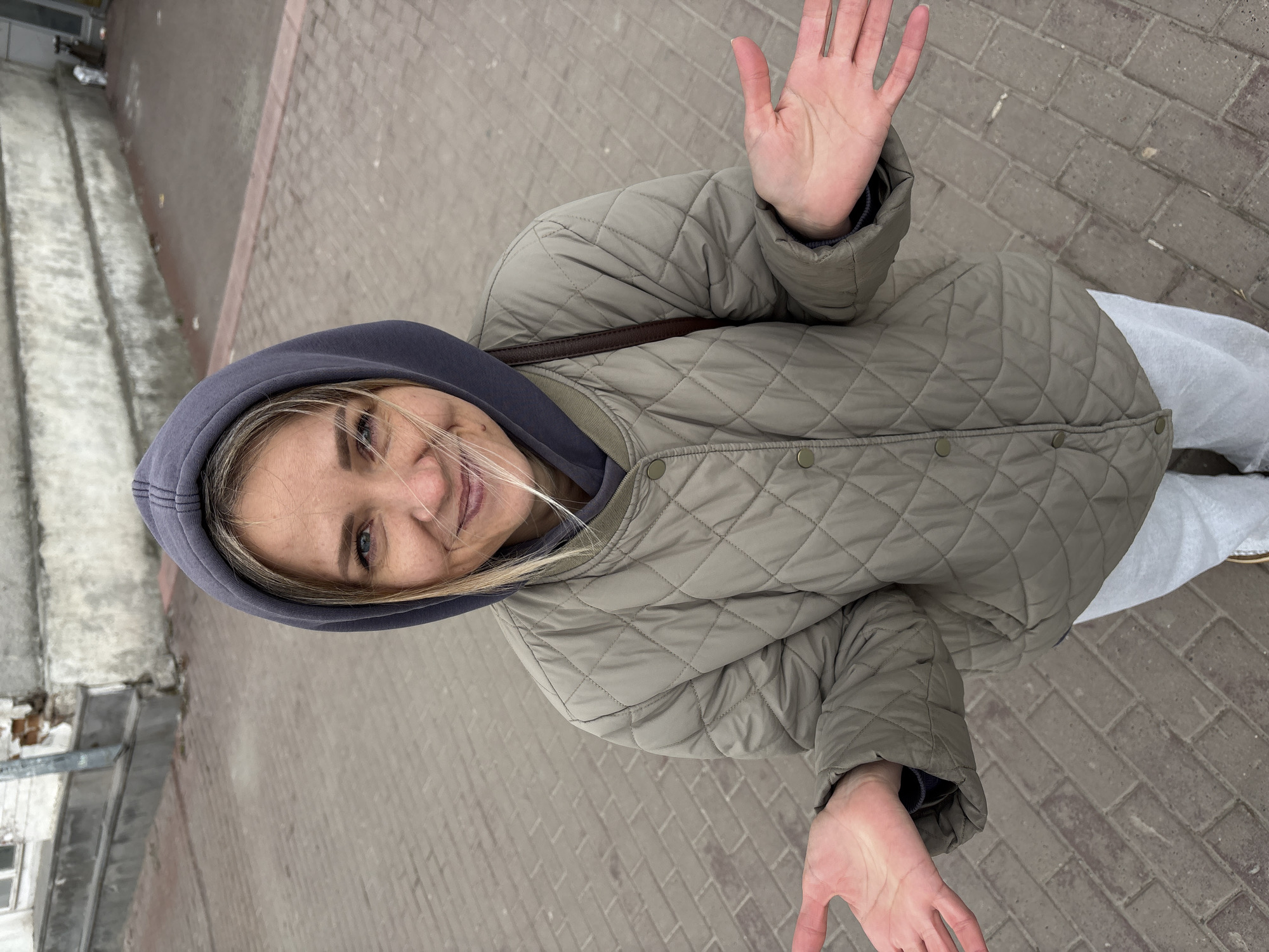 A woman is standing on a brick-paved surface in Ulyanovsk, Russia. She is wearing a hooded sweatshirt with the hood up, and a quilted jacket. She is facing the camera with both hands raised, palms open. The location appears to be a street or pedestrian area. Buildings are visible in the background, with a light-colored structure on the left side of the frame.