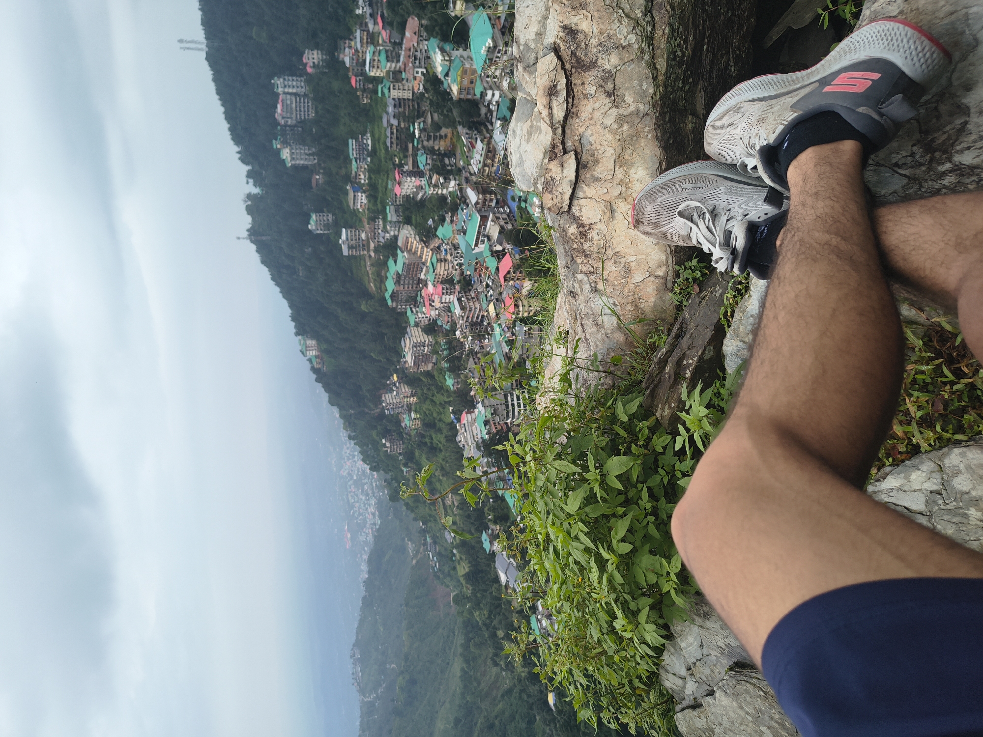 A first-person perspective shot captures a person's legs and feet resting on a rocky outcrop overlooking the city of Bhagsu, India. The person is wearing dark blue shorts and grey athletic shoes. The foreground is dominated by the rocky terrain and some lush green foliage. In the midground and background, the hillside is densely populated with buildings, many featuring colorful roofs, and is covered in trees. The city sprawls down the mountainside, with the distant landscape fading into a hazy, overcast sky. The overall scene depicts a moment of rest and contemplation with a panoramic view of the urban and natural environment.