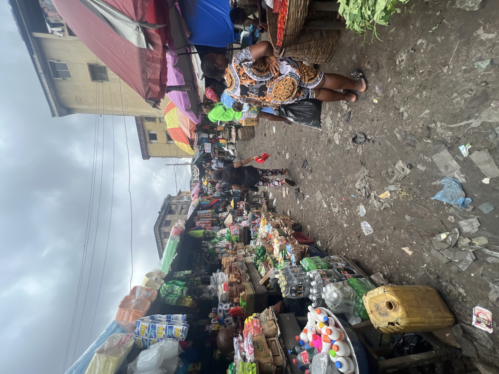 This is a ground-level, eye-level photograph of a crowded outdoor market in Nkpor, Nigeria. The scene depicts a vibrant but cluttered environment with numerous stalls selling various goods, including food items, drinks, and household products. People are actively engaged in shopping and selling, with some individuals carrying bags and others tending to their stalls. The ground is covered in debris, suggesting a busy and well-trafficked market. The overall impression is one of a bustling commercial hub, typical of urban Nigerian settings.
