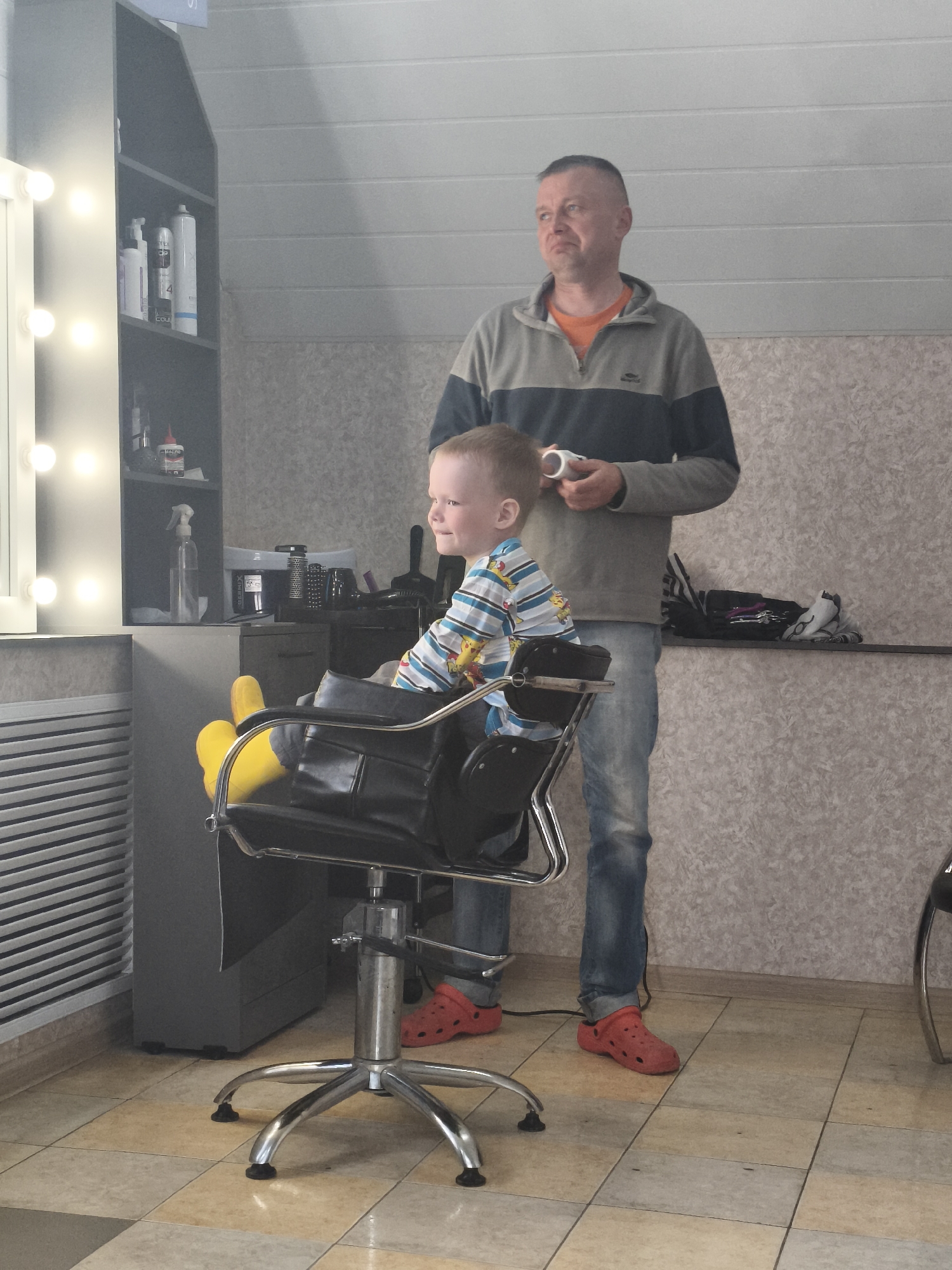 A young boy sits in a black salon chair with bright yellow supports under his feet. The boy wears a blue and white striped shirt. A middle-aged man stands behind him, holding a white cup. The man wears jeans, a gray and blue striped sweater, and red Crocs-style shoes. They are in a room with tiled floors, and a gray cabinet with beauty supplies is visible to the left of the boy. The location is Русский Вожой, Russia. A counter with various salon implements is visible in the background.