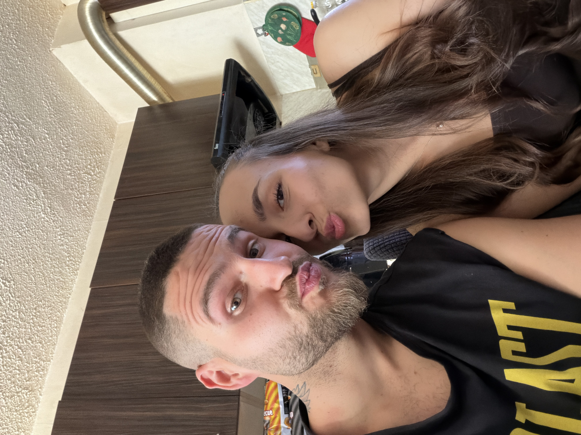 A selfie depicts a man and a woman, their faces close together, both pouting their lips toward the camera. The man has fair skin, a short haircut, a beard, and is wearing a black shirt with yellow lettering. The woman has long, brown hair and is wearing a black top. The background reveals a kitchen area with light-colored walls and dark brown cabinets. Other kitchen objects such as a range hood, and a package of chips are visible. The image was likely taken indoors. The location (Sofia, Bulgaria) cannot be verified based on the visual information provided.
