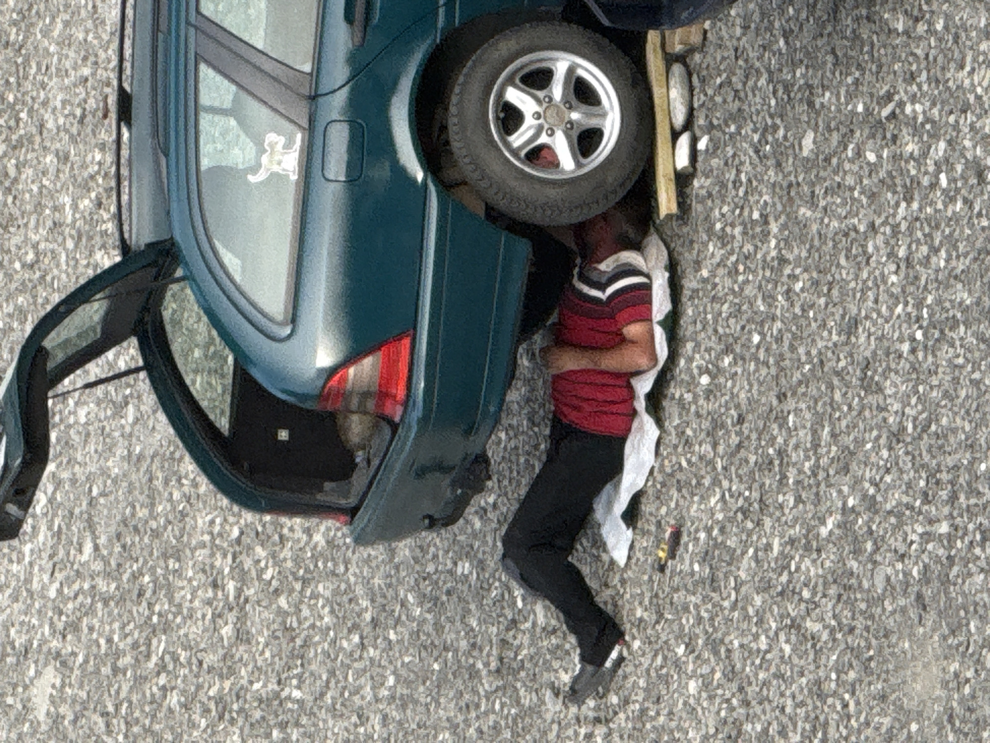 In Tbilisi, Georgia, a person is lying on their back on the ground, underneath the rear of a dark green station wagon. The trunk of the car is open, revealing the interior. The person is wearing a red and white striped shirt and black pants. They are lying on a white cloth or mat, with tools such as a screwdriver and pliers nearby. The ground is covered in gravel. The overall scene suggests car repair or maintenance is taking place outdoors.