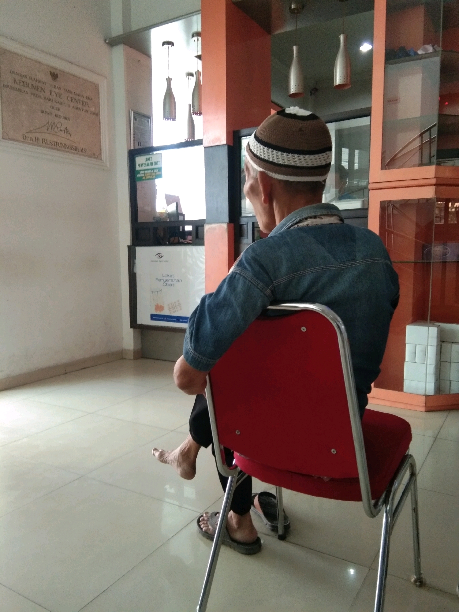 An individual, wearing a brown and white patterned cap and a blue denim jacket, sits on a red chair in what appears to be a waiting area. The individual is facing away from the camera, with their left foot extended and bare, while their right foot is wearing a grey sandal. In the background, a framed document is mounted on a white wall, with text in Indonesian that includes "DENGAN RAHMAT TUHAN YANG MAHA ESA KEBUMEN EYE CENTER" and mentions "Dra. Hj. RUSTIRINGSIH, M.SI". There is also a sign above a counter with the text "LOKET PENYERAHAN OBAT". The environment features tiled flooring and an orange-colored partition with glass display cabinets. The overall scene suggests a medical facility or clinic in Kebumen, Indonesia.