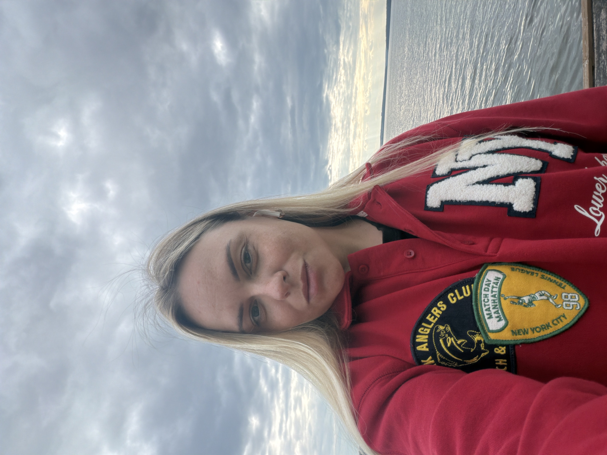 A selfie taken from a woman's chest up shows her from the chest up, facing the camera, with a body of water and cloudy sky in the background. The woman has blonde hair and is wearing a red jacket with patches and embroidery. The patches include a fish in a circle and a tennis player with the text "MATCH DAY MANHATTAN NEW YORK CITY TENNIS LEAGUE 98". There is also an embroidered "N" on the jacket. A white wireless earbud is visible in her left ear. The scene is set outdoors, likely near a body of water, with overcast skies. No specific landmarks or details definitively place the location in Votkinsk, Russia.