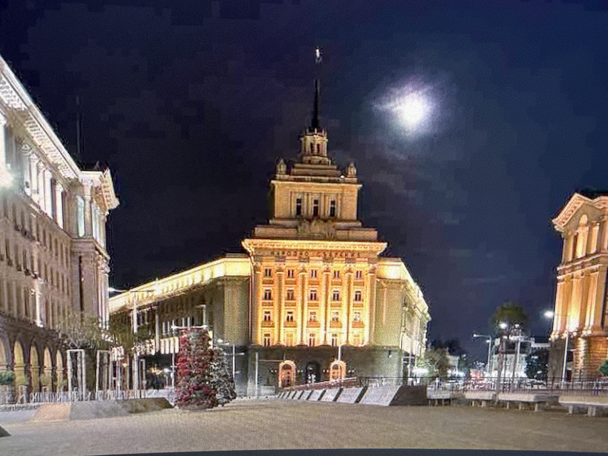 The image shows a night view of a building illuminated with golden light. It is the Largo in Sofia, Bulgaria, likely taken from Independence Square. The architecture is Soviet-era, and a Christmas tree stands near the building's entrance. A bright moon is visible in the dark sky above. The area in the foreground is a large open plaza. Buildings flank the main structure on either side. Street lamps are scattered around the plaza. There are no people clearly visible.