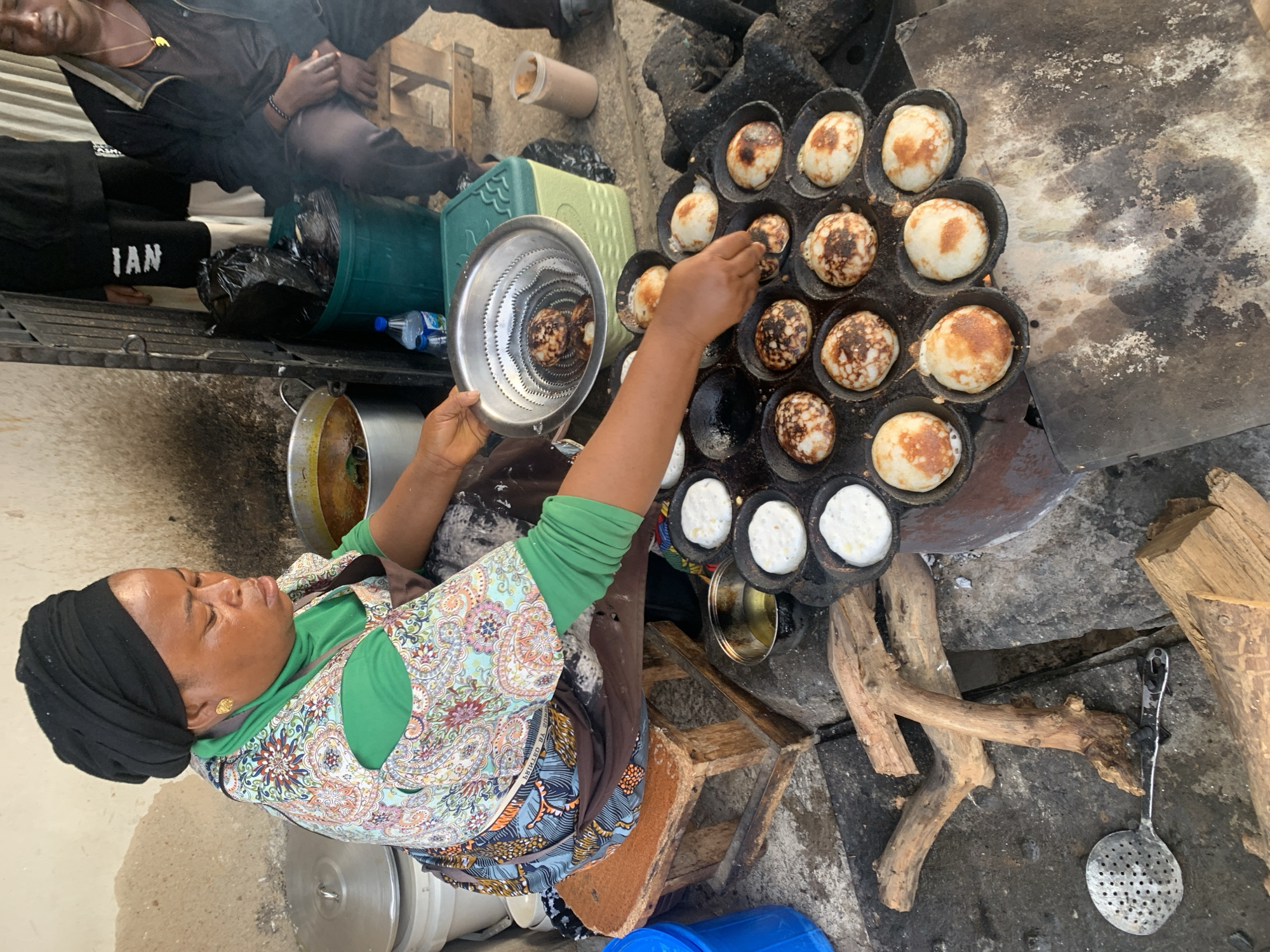 In Jos, Nigeria, a woman wearing a black headscarf and a colorful patterned dress is cooking small, round pancakes in a traditional open-air cooking setup. She is seated on a small wooden stool next to a cast-iron pan with multiple circular molds, where the pancakes are being cooked over what appears to be charcoal embers. The woman is actively involved in the cooking process, holding a metal strainer containing some cooked pancakes and using her right hand to adjust another pancake in the mold. Beside her, a pot containing a stew or soup is visible. In the background, other individuals are present, one of whom is seated and another standing. The environment appears to be an outdoor marketplace or street vendor stall, with various containers, bags, and other cooking utensils scattered around. The overall scene depicts a common street food preparation process in Nigeria.