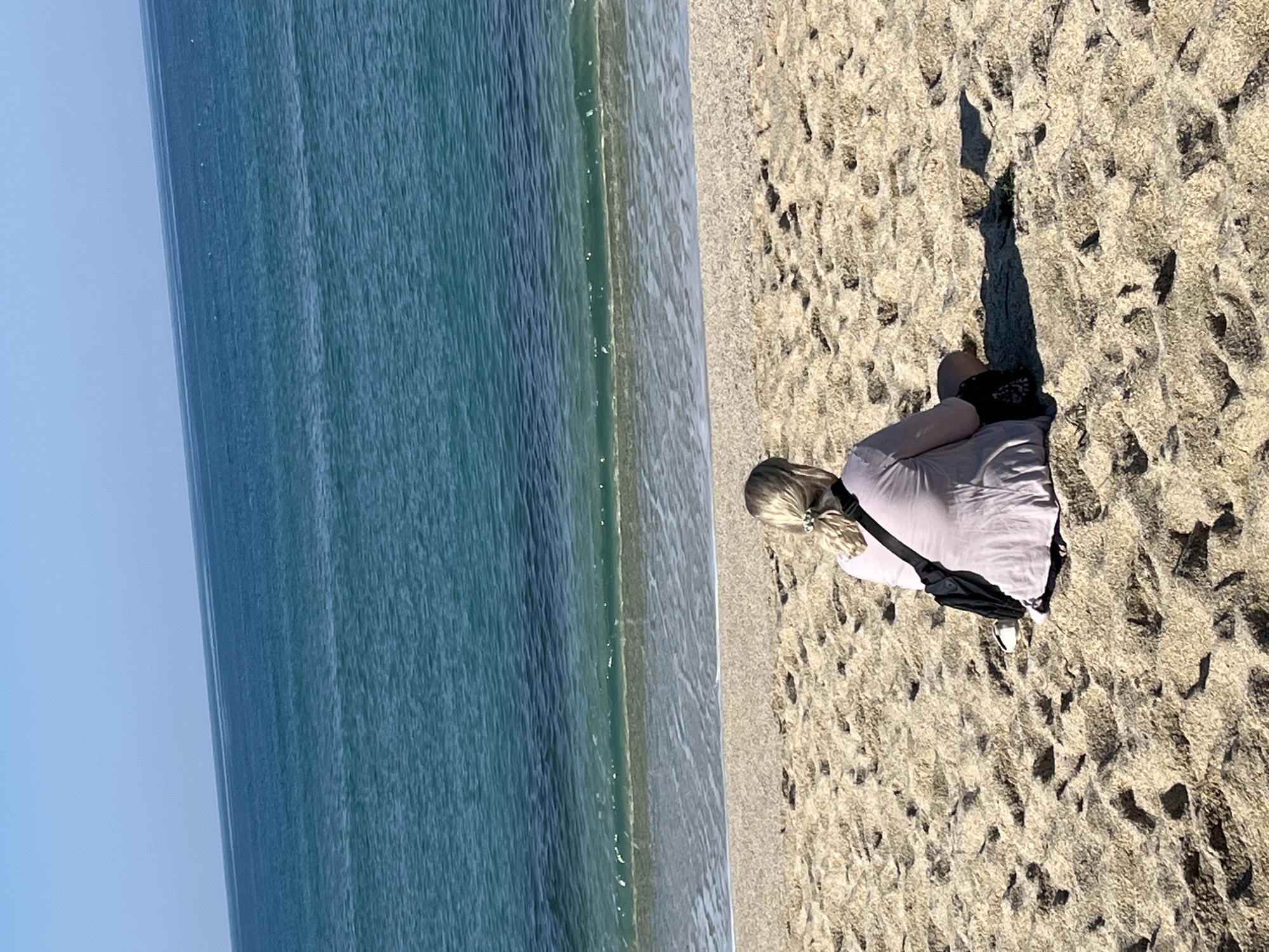 A high-angle photograph shows a woman sitting on a beach in Alanya, Turkey. The woman has blond hair pulled back with a hair tie. She is wearing a light purple shirt, black pants, and white shoes, with a black bag across her chest. She sits on sand next to the turquoise ocean. The sky is clear and blue. The sand has uneven patterns and dark spots. The water is clear close to shore and deeper blue further away from the shore.