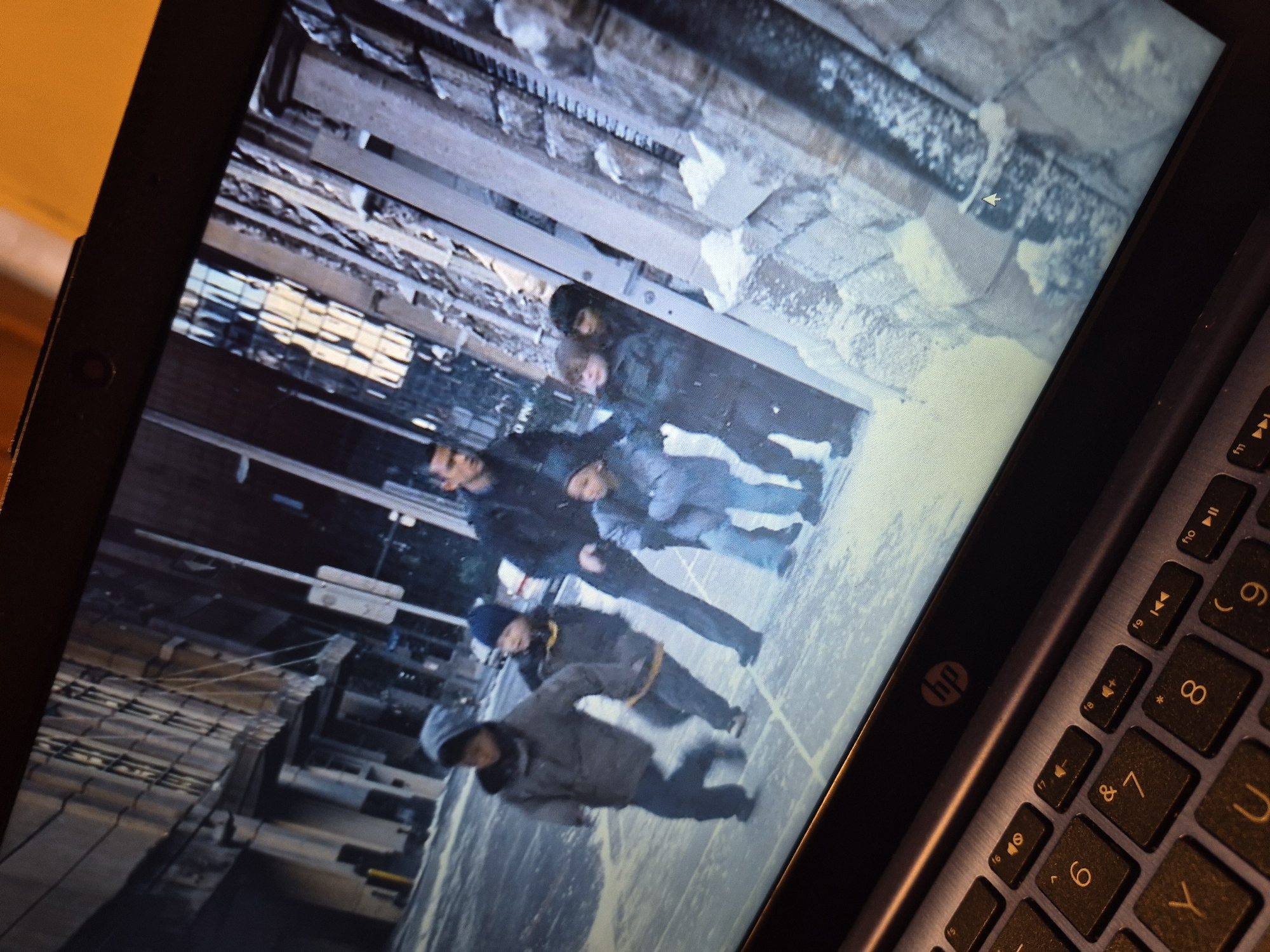 The image displays a laptop screen showing a scene from a movie or TV show. The scene appears to be set in a snowy urban environment, with several children and an adult walking in what looks like a street. The buildings in the background are tall and appear to be made of concrete and glass. There is snow on the ground and on the sides of the buildings. The people are dressed in winter clothing, with the children wearing hats and gloves. The adult appears to be leading the children. The laptop itself is shown in the foreground, with its keyboard visible. The HP logo is visible on the laptop's bezel. The image does not contain any information that would allow for the identification of the location as Sandwell, United Kingdom.