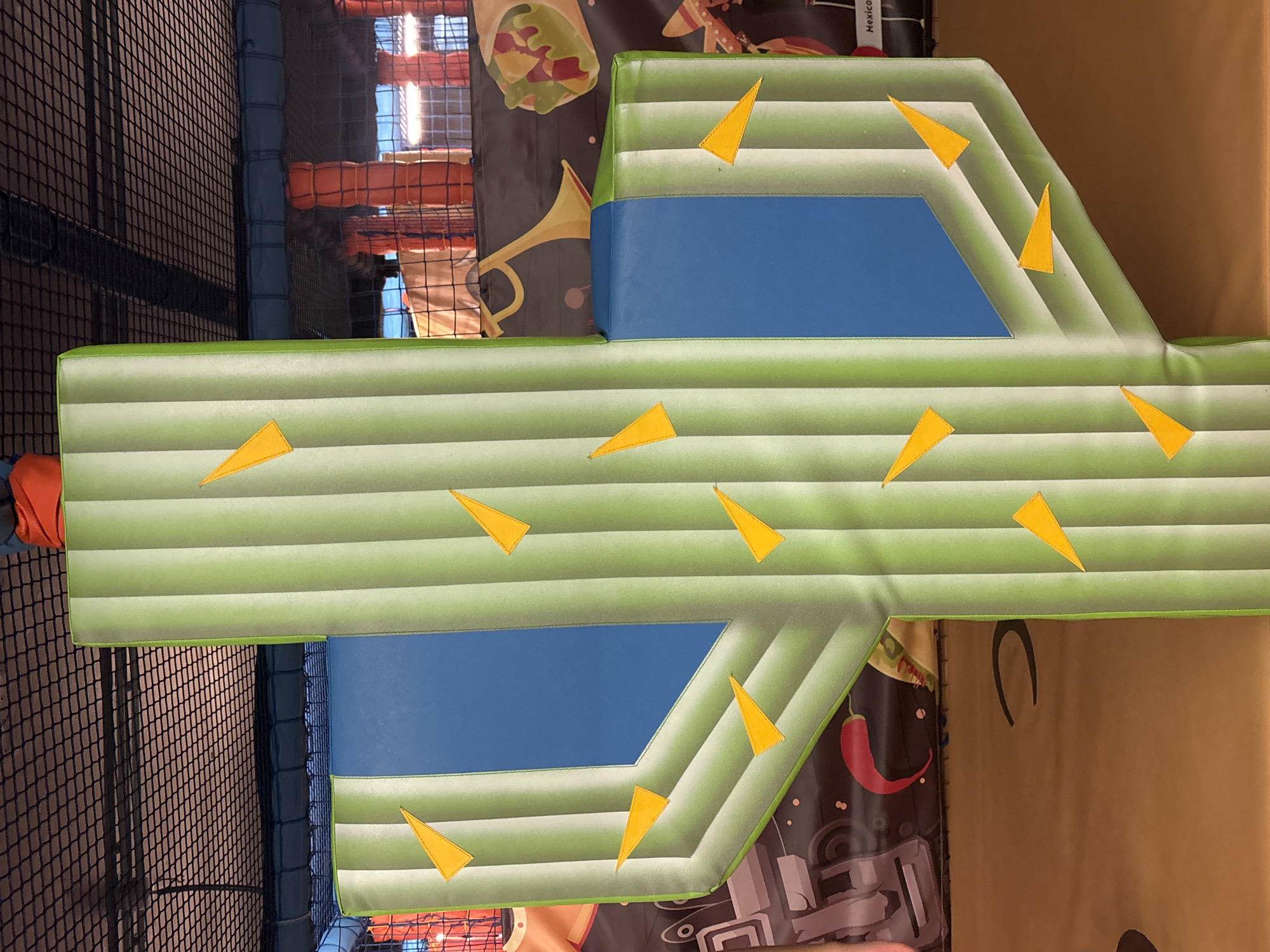 The image shows a brightly colored, inflatable cactus structure with blue inserts, decorated with yellow triangular elements. The cactus appears to be part of a play area, possibly an indoor playground, as suggested by the netting and padded flooring visible in the background. The overall theme seems to be festive or themed, with hints of Mexican motifs in the surrounding decorations, such as chili peppers and what appears to be a burrito. The lighting is artificial and casts shadows, indicating an indoor environment. There are no people visible in the frame. The exact location in Dubai, United Arab Emirates, is not discernible from the image itself.