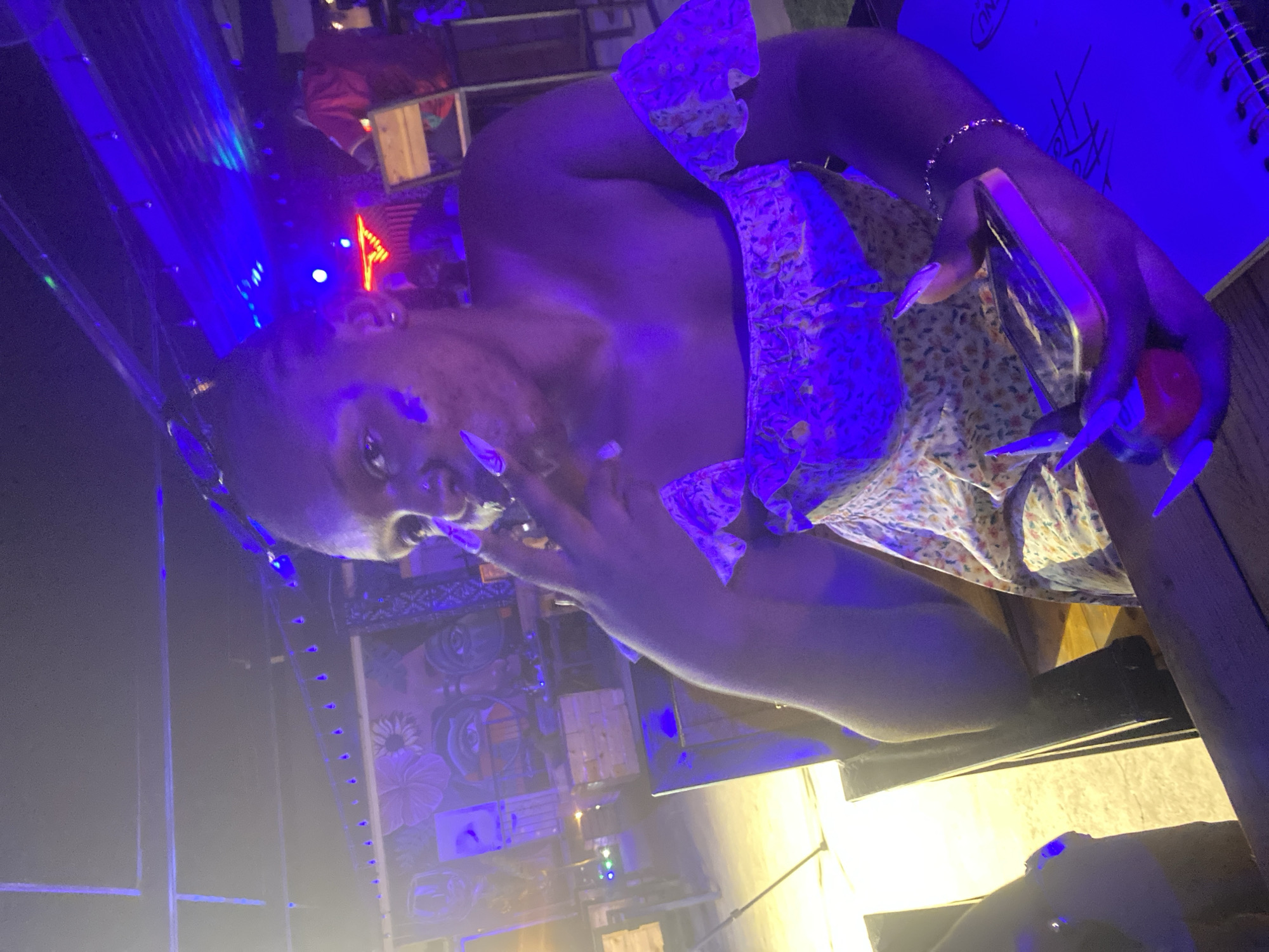 The image shows a person with dark skin, adorned with long, pointed, lavender-colored fingernails, wearing a floral-patterned dress. The person is holding a red object, possibly a small toy or fruit, in their right hand. Their left hand is raised towards their face. The scene is dimly lit, with vibrant blue and yellow lights illuminating the surroundings. In the background, there are some abstract murals on a wall and what appears to be a notebook or planner with some scribbles on it. The lighting suggests it might be nighttime or an indoor setting with artificial lighting. The person's expression is difficult to discern due to the lighting and angle. The image seems to capture a moment of personal expression or enjoyment, possibly at a party or social gathering, given the ambiance of the lighting and the presence of what could be decorative elements. There is no visible text that can be clearly read. The location is identified as Howayi, Nigeria.