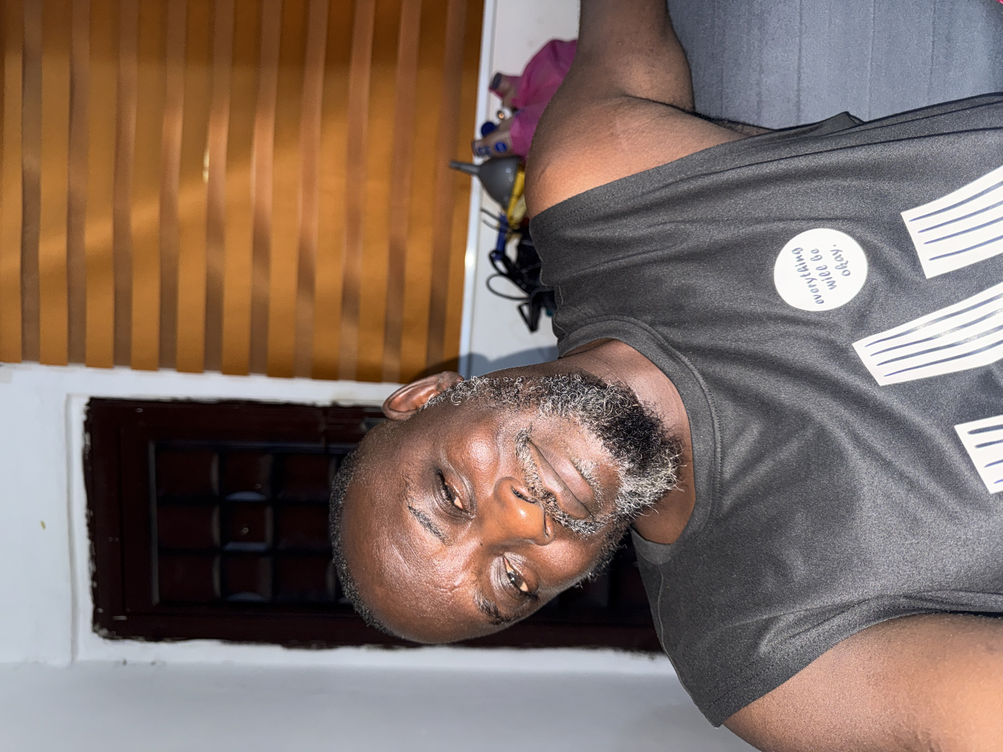 A middle-aged Black man with a graying beard is shown in a close-up shot. He is wearing a dark gray t-shirt with a white circular design and horizontal white stripes. He appears to be indoors, against a wall with brown horizontal blinds. To the left of the man is a dark brown window with a grid pattern. Based on the prompt, it is assumed the location is in the city of Ado, Nigeria.