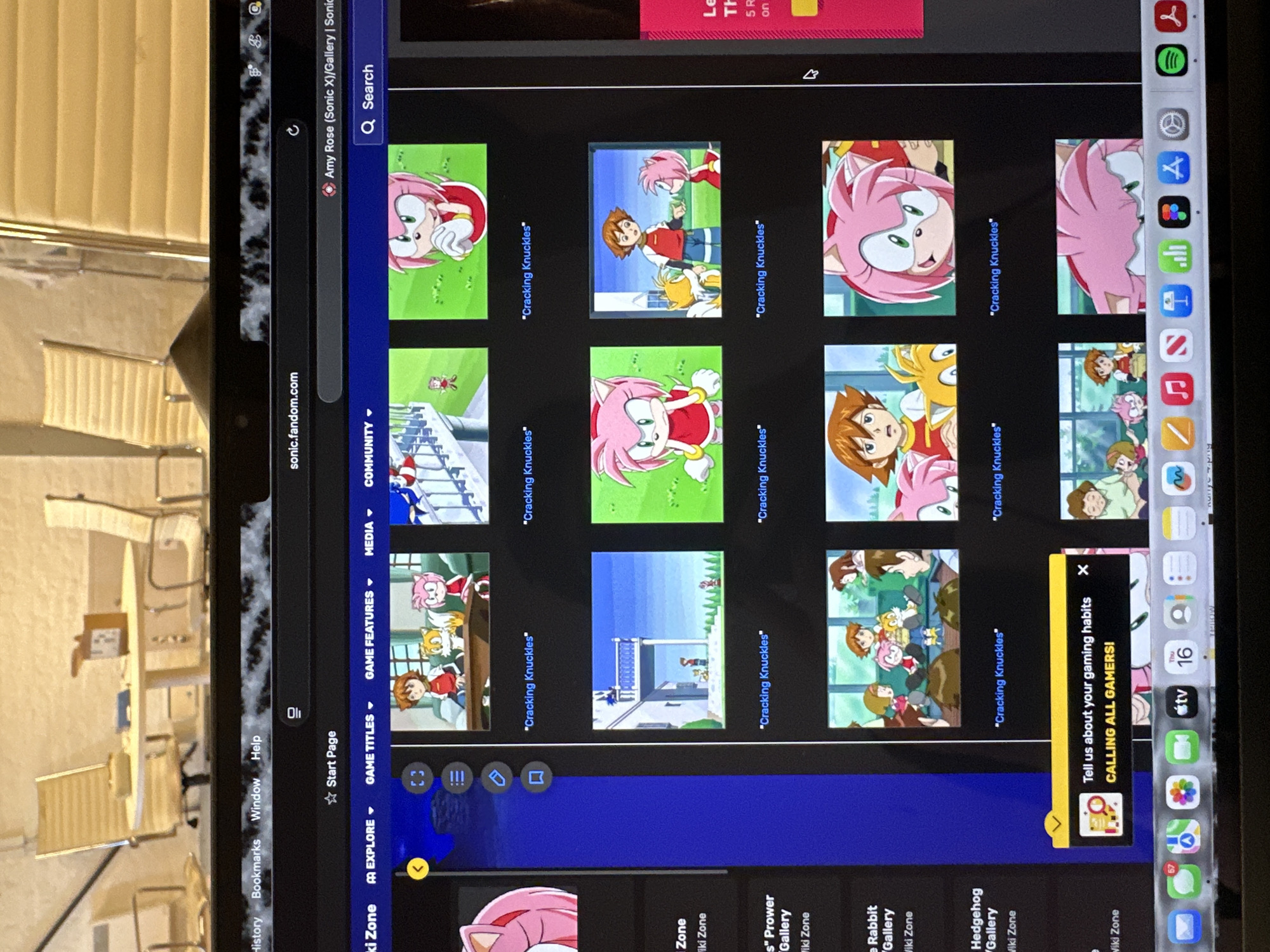 An electronic tablet displays a webpage from sonic.fandom.com, showing a gallery for "Amy Rose (Sonic X)." The tablet is oriented horizontally, with its screen visible from an overhead angle, while a blurred background reveals light-colored chairs and a table in an indoor setting.
The main activity is internet browsing, specifically viewing animated character images from the Sonic X series. The prominent character, Amy Rose, a pink hedgehog, is featured in many of the thumbnail images, often alongside other characters like Chris Thorndyke and Tails. Several image captions repeat the phrase ""Cracking Knuckles"".
Notable details include the website's URL (sonic.fandom.com) and the clear page title, "Amy Rose (Sonic X)/Gallery | Sonic". The browser interface shows various navigation options like "History," "Bookmarks," "Window," "Help," and categories like "EXPLORE," "GAME TITLES," "GAME FEATURES," "MEDIA," and "COMMUNITY." A search bar is also visible. On the right side of the screen, an orange-yellow pop-up banner advertises, "Tell us about your gaming habits CALLING ALL GAMERS!". The tablet's status bar shows "Thu 16" and a battery indicator. A vertical dock or sidebar on the far right displays several application icons, including Spotify, Safari, App Store, Files, Photos, and an Apple TV app, with a messages app showing a notification badge of "67". The background, although out of focus, suggests a contemporary indoor environment, possibly a home or cafe setting, with soft lighting typical of daytime. The image content does not provide specific cues to confirm the London, UK location.