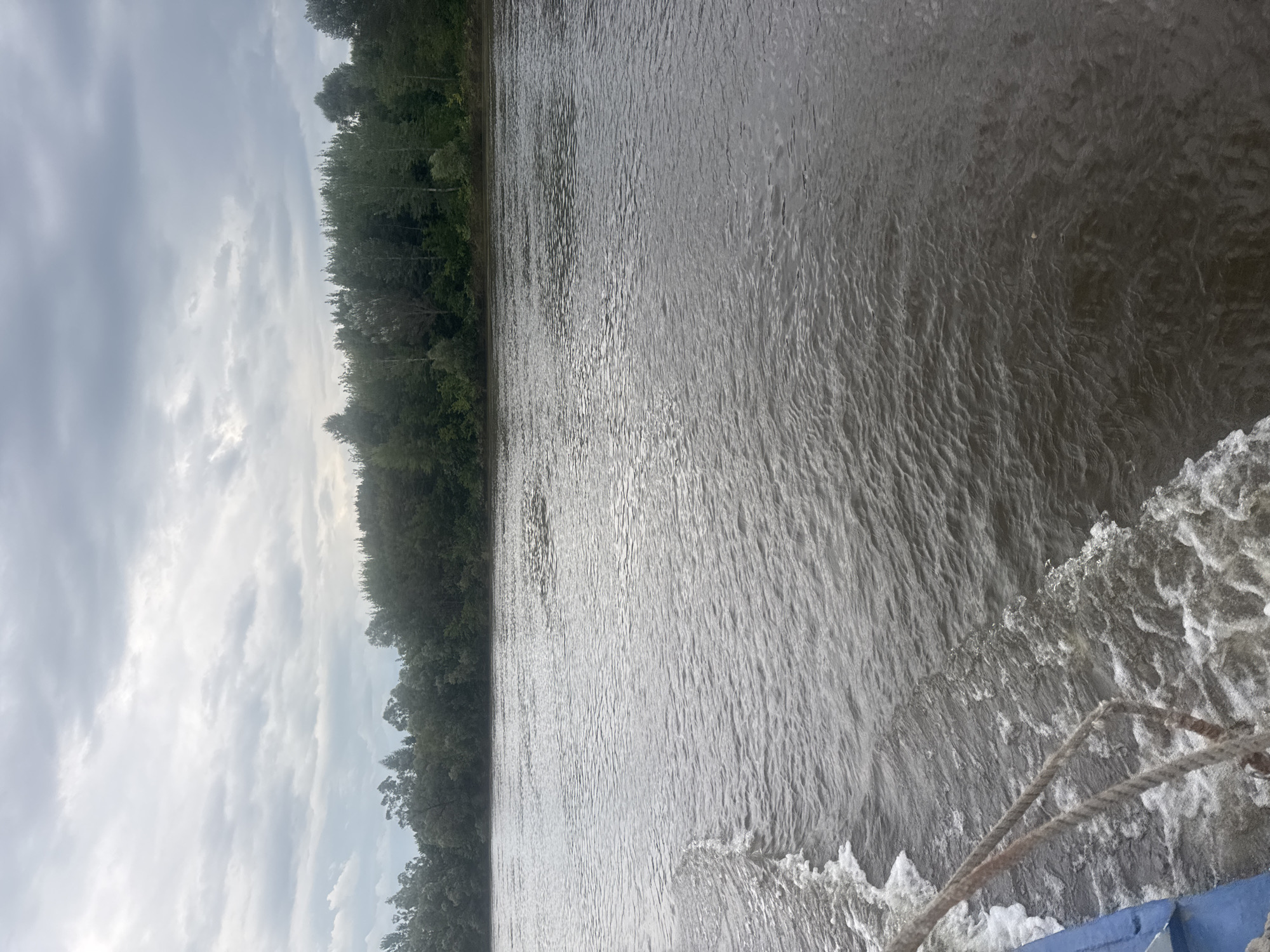 A view from a boat moving on a wide, murky river, with a dense forest lining the distant bank under a cloudy sky. The river water is choppy, with white foam trailing behind the boat, indicating movement. A rope is visible in the foreground, attached to the blue side of the boat. The scene is likely the Ufa River in Ufa, Russia, given the presence of a wide river and a thick forest, characteristic of the region's natural landscape.