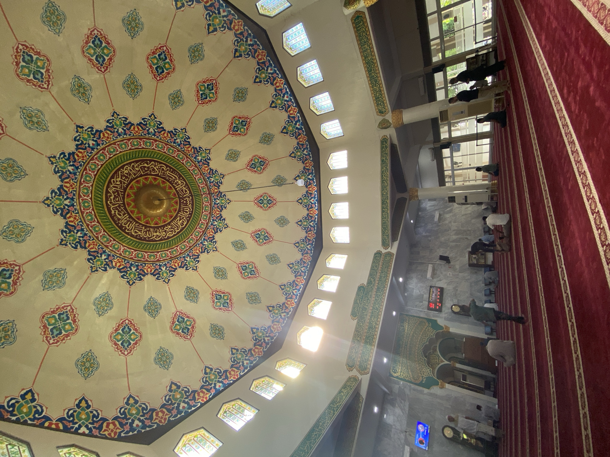 The image shows the interior of the Islamic Center Lhokseumawe mosque in Lhokseumawe, Indonesia. The main focus is on the ornate ceiling featuring a large, circular design with intricate patterns, calligraphy, and a central golden dome. The ceiling's color scheme includes blues, greens, reds, and cream tones. Windows are aligned along the perimeter of the ceiling. Along one side is a red carpet that stretches the length of the prayer hall. Worshippers are present, some standing, kneeling, or prostrating in prayer. The environment is brightly lit from natural and artificial lighting, with columns featuring inscriptions. An elaborately decorated mihrab (niche in the wall indicating the direction of Mecca) is also visible. A digital clock is mounted on the wall.