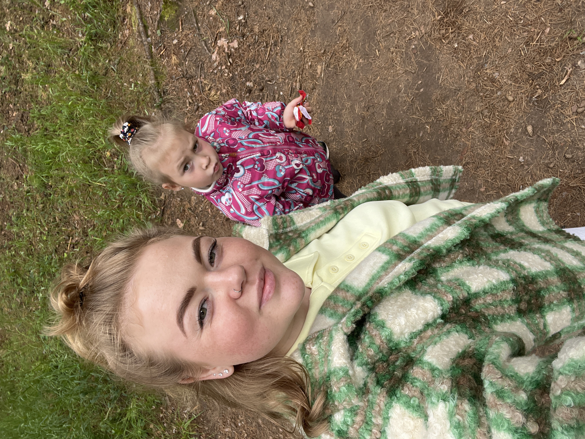 This image depicts a young adult woman and a young child in an outdoor setting in Chaikovsky, Russia. The woman, positioned in the foreground and taking a selfie, has light hair pulled into a messy bun, fair skin with freckles, light-colored eyes, a nose ring on her left nostril, and three stud earrings on her left ear. She is wearing a light yellow collared shirt beneath a green, white, and brown plaid fuzzy jacket, and is looking directly at the camera with a subtle smile.
Behind her and to the right, a young child stands, looking towards the camera with a slight frown or pout. The child has light hair secured in a high ponytail with a colorful scrunchie and is wearing a pink jacket adorned with a repeating pattern of white and blue key-like shapes. The child holds a crumpled red and white wrapper in their right hand.
The ground consists of dark brown soil covered with an abundance of dried pine needles, small twigs, and scattered pinecones, indicative of a forest floor. Patches of vibrant green grass are visible towards the upper left and central left portions of the background. The lighting appears natural and diffused.