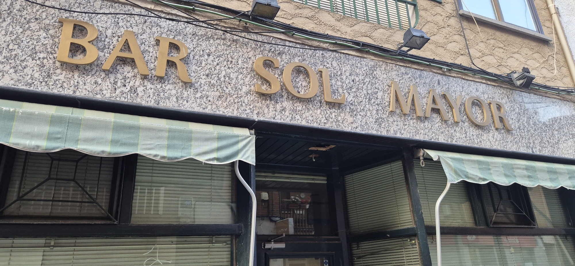 The image shows the facade of a bar named "BAR SOL MAYOR" in a city. The bar's name is displayed in large, gold-colored, three-dimensional letters mounted on a speckled granite-like facade. Below the signage, there is an awning with green and white stripes, and a dark-framed storefront with windows covered by blinds. Electrical wires and lighting fixtures are visible above the signage, attached to the textured exterior wall of the building. The overall impression is of a commercial establishment in an urban setting.